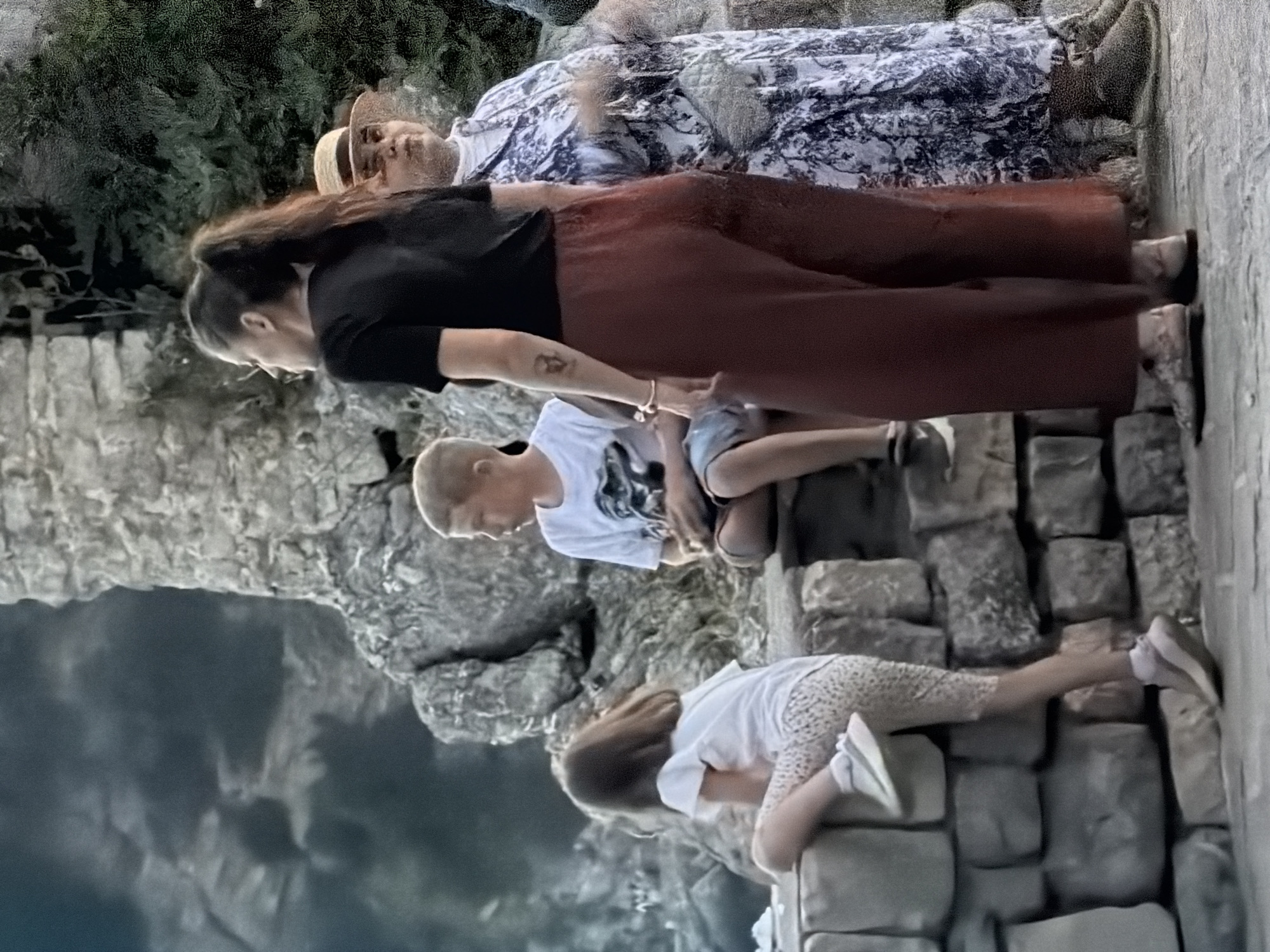 In Budva, Montenegro, a woman with a dark ponytail wearing a black t-shirt and rust-colored wide-leg pants stands facing away from the camera. To her left, a young boy in a white t-shirt with a graphic print is seated on a stone wall, looking to his right. A young girl with brown hair, wearing a white t-shirt and patterned leggings, is climbing on the same stone wall, facing away from the camera and looking to her left. To the right of the woman, an older woman in a blue and white floral dress and a straw hat looks to her left. The environment appears to be outdoors, with a background of textured stone structures and lush green foliage. The lighting suggests it is either late afternoon or early evening.