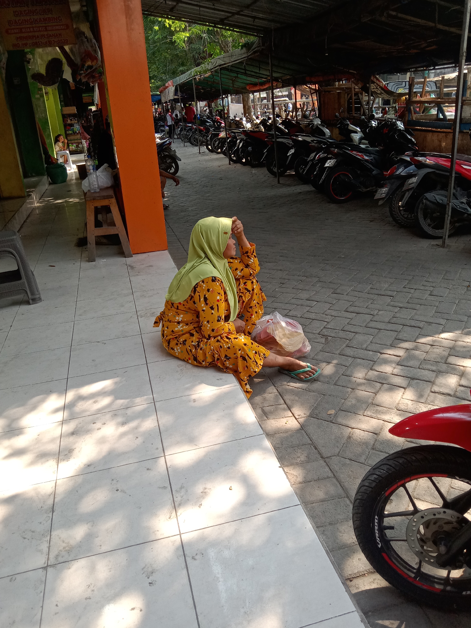 The image captures an outdoor scene likely in Sugihwaras, Indonesia, characterized by a tiled walkway adjacent to a cobbled surface and a covered area with parked motorcycles. A woman wearing a light green hijab and a yellow patterned dress is seated on the tiled ground, her body angled away from the camera. She appears to be resting her forehead on her hand. In front of her, a plastic bag containing what appears to be food items rests on the ground, along with a pair of green flip-flops. The background reveals a row of motorcycles parked under a canopy, suggesting a marketplace or public area. Sunlight casts dappled shadows across the tiled surface. A red motorcycle fender and wheel are visible in the foreground on the right. The environment suggests a warm, sunny day.