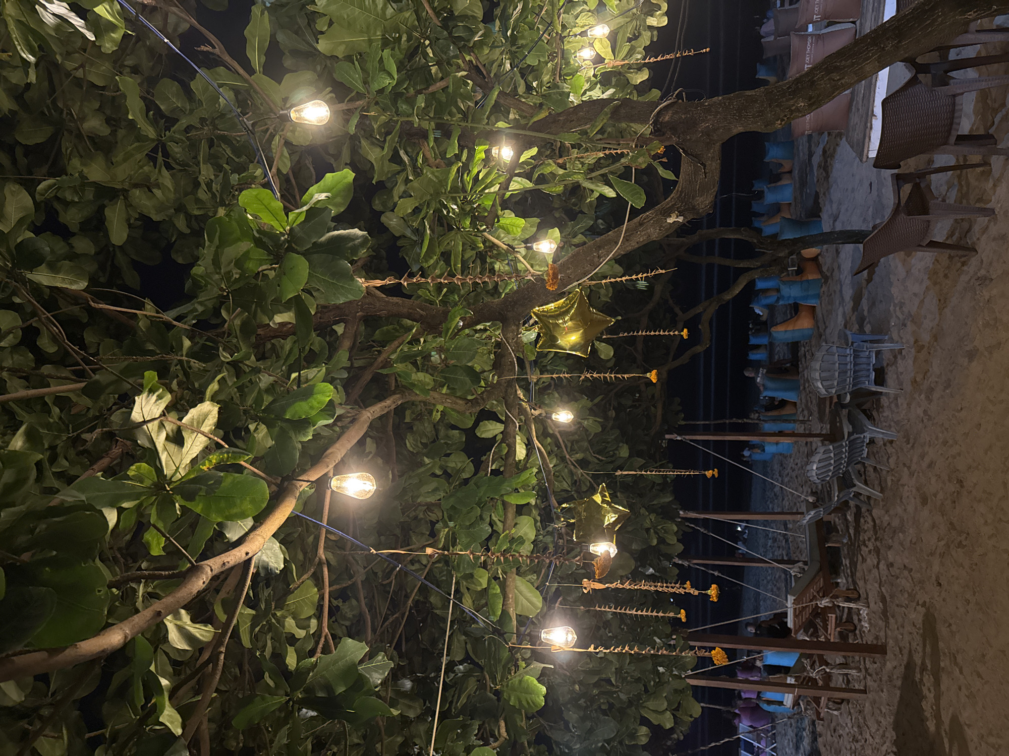 The image depicts a tropical beach scene at night in Kuta, Indonesia. The foreground is dominated by a large tree with dense green foliage, from which numerous string lights are hung, casting a warm glow. The branches of the tree are adorned with decorative strings of small orange flowers and what appear to be dried seed pods, along with several shiny gold star-shaped balloons. Beneath the tree, on the sandy beach, are several lounge chairs and small tables, some with chairs, suggesting an outdoor seating area. In the background, more beanbag-style seating in blue and orange hues is visible, arranged on the sand. The ocean is faintly visible in the distance, with the dark expanse of the night sky above. The scene appears to be set up for evening relaxation or dining at a beachside establishment.