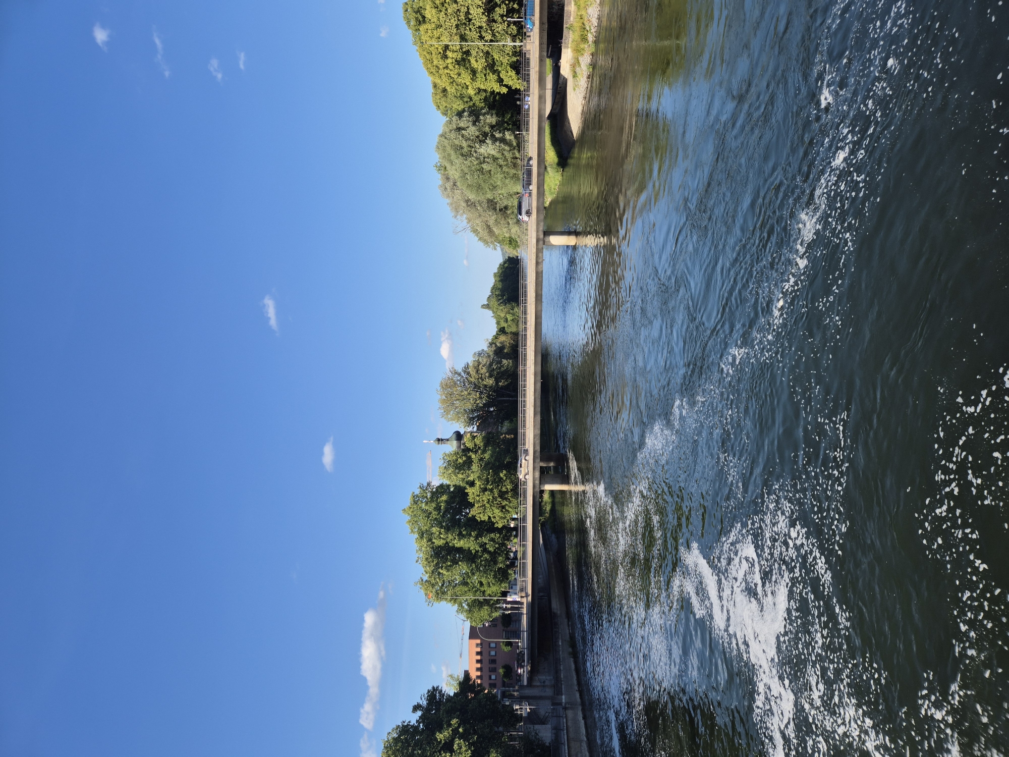 The image depicts a wide river flowing through the city of Landshut, Germany. A concrete bridge spans the river, carrying vehicular and pedestrian traffic. On the left bank, a multi-story brick building is visible, partially obscured by trees. A church spire with a green dome rises above the trees in the background. The right bank is lined with lush green trees. The river's surface is a mixture of dark water and frothy white foam, indicating a current. The sky is a clear, vibrant blue with a few scattered white clouds. The lighting suggests it is a sunny day, likely late morning or early afternoon. There are several cars and a few people visible on the bridge.
