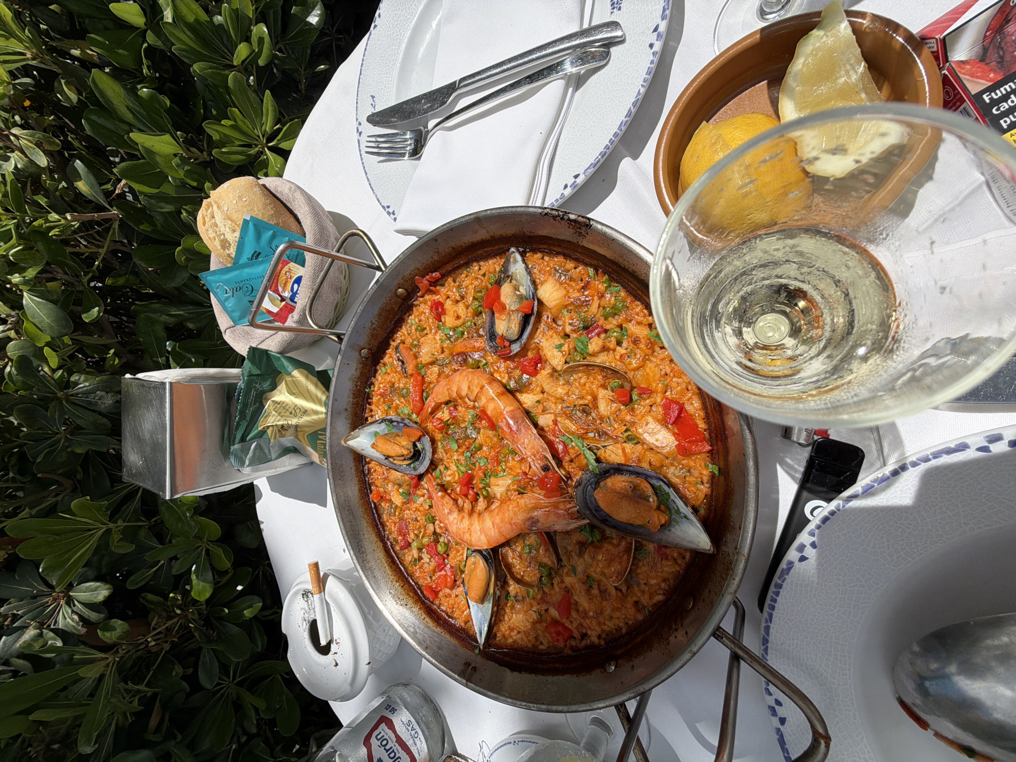 The image captures a table setting in Benalmádena, Spain, featuring a paella dish as the main subject. The paella, cooked in a large, shallow pan, contains seafood such as shrimp and mussels, along with rice and vegetables. Adjacent to the paella are a wine glass filled with white wine, a bowl of lemon wedges, and a basket of bread with condiment packets. Cutlery and plates are neatly arranged on the table. Next to the paella, there's an ashtray with a cigarette. Also present are a lighter, a butane gas bottle for portable stoves, and a pack of cigarettes. The table is positioned outdoors next to some greenery.