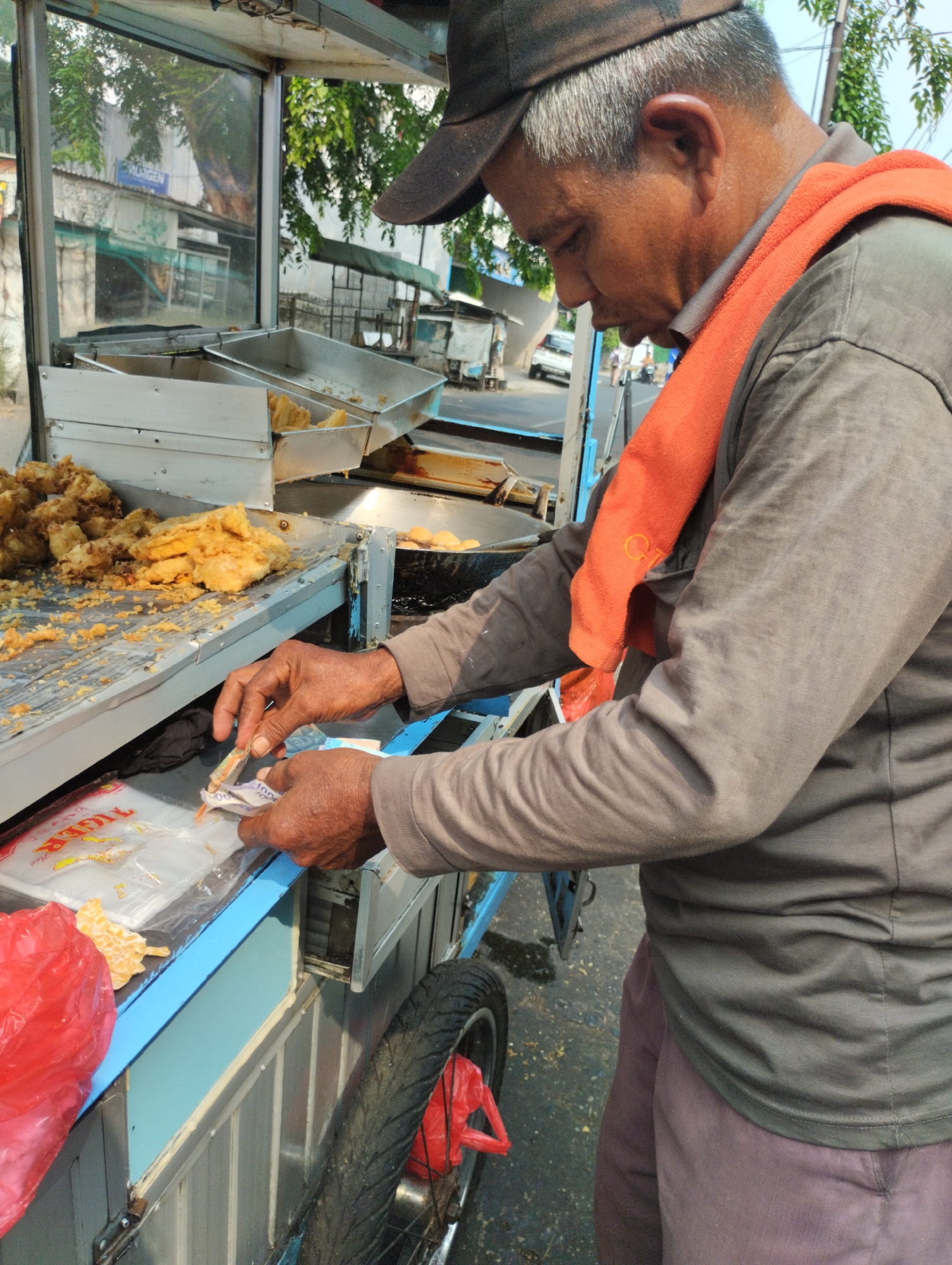 A street food vendor, an Indonesian man wearing a dark baseball cap and a grey long-sleeved shirt with an orange towel draped over his shoulders, is seen from the side and slightly from behind, operating a food cart in Bekasi, Indonesia. He is holding Indonesian currency, a few bills in his hands, and appears to be handling a transaction. The food cart is primarily made of metal and glass, with a blue trim along the lower section. Visible inside the cart are containers filled with fried food items, likely snacks or finger foods, and a deep fryer in the background. Adjacent to the cart, a red plastic bag and a portion of a wheel are also present. The background reveals a street scene with buildings and trees, typical of an urban environment in Indonesia.