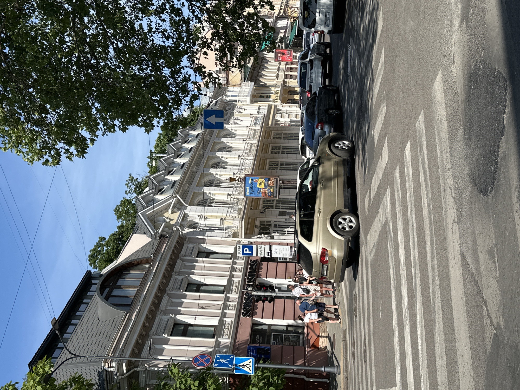 This forensic image captures a sunny daytime street scene in Odesa, Odesa Oblast, Ukraine. The perspective is from a slight elevation, looking down a city street lined with ornate multi-story buildings, characteristic of late 19th/early 20th-century architecture.
In the immediate foreground, a faded zebra pedestrian crossing is visible on the asphalt pavement. A gold-colored SsangYong Kyron SUV (license plate BH 0023 MX) is positioned on the right side of the road, partially obscuring the view of the street ahead, with its rear facing the camera. The asphalt shows signs of wear and patching.
On the left sidewalk, a group of five pedestrians – three males and two females, all appearing to be adults in casual summer attire – are standing near a lamppost with multiple traffic signs. These signs include a "no stopping" sign, a "pedestrian path" sign, and a "pedestrian crossing ahead" sign. A red pedestrian signal is visible on a pole in front of a building that houses "SENSE BUSINESS" on its ground floor. This building features a light pink/beige facade with neoclassical architectural details and a dark grey mansard roof with an arched dormer window.
Further down the street, a line of vehicles is queued in traffic, facing away from the camera. Visible vehicles include a dark sedan, a white SUV, and a white light commercial vehicle. A blue "straight ahead only" directional sign is mounted above the street. On the right side of the street, more historical buildings with balconies and decorative elements are visible. Advertising banners hang from these buildings, including one for "lifecell" (a Ukrainian mobile operator) and a red banner with the Ukrainian word "СУКНЯ" (dress).
Lush green trees are prominent on the right side of the image, casting shadows on the street, and more trees are visible atop a hill behind the buildings on the left. The sky is clear and blue, indicating bright sunlight. A parking zone sign ("ЗОНА P") with a bus stop symbol and numeric information ("10 25.50" and "30") is visible on the right sidewalk near the "SENSE BUSINESS" building.