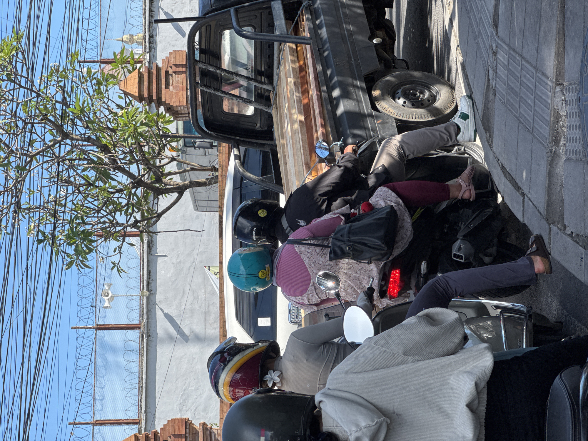 The image captures a sunny street scene in North Kuta, Indonesia, featuring several individuals on motorbikes and a pickup truck, likely stopped in traffic or parked. The time of day appears to be midday or early afternoon, indicated by the bright, clear blue sky and strong shadows.
In the foreground, the backs of multiple people on scooters or motorbikes are visible. One individual wears a black helmet with a yellow sticker, another a teal helmet with "YNA" visible, and a third a red and yellow helmet with a white frangipani flower tucked behind their ear. People are dressed casually, with some wearing long sleeves and pants, possibly for sun protection. Footwear visible includes light pink sandals, white and green sneakers, and brown loafers. "HONDA" is visible on one of the helmets.
Behind the motorbikes, a dark-colored pickup truck with a wooden or metal flatbed structure is present. To the left, a frangipani tree with green leaves and white blossoms frames the upper portion of the scene. In the background, part of a light-colored building with a gray wall is visible, along with a reddish-brown brick structure resembling a traditional Balinese shrine or architectural element. Numerous overhead electrical or communication wires crisscross the sky. A fence or wall topped with barbed wire is also discernible. The overall atmosphere is that of a bustling street in a tropical environment.