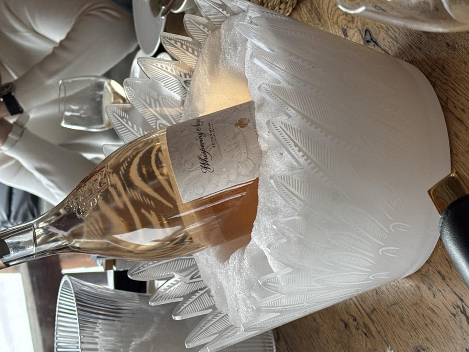 A bottle of "Whispering Angel 20th Vintage Limited Edition" rosé wine stands upright within a decorative, frosted white ice bucket filled with granular ice, situated on a light-colored wooden table. The ice bucket features an intricate, etched pattern resembling stylized feathers or leaves across its surface and rim. In the mid-ground, a person wearing a white long-sleeved garment is partially visible, with hands clasped and a watch on their left wrist. A stemmed wine glass, partially filled with rosé wine, is visible to the right of the ice bucket, and a ribbed clear glass carafe is present to its left. The scene depicts a dining or social setting in Courchevel, France.