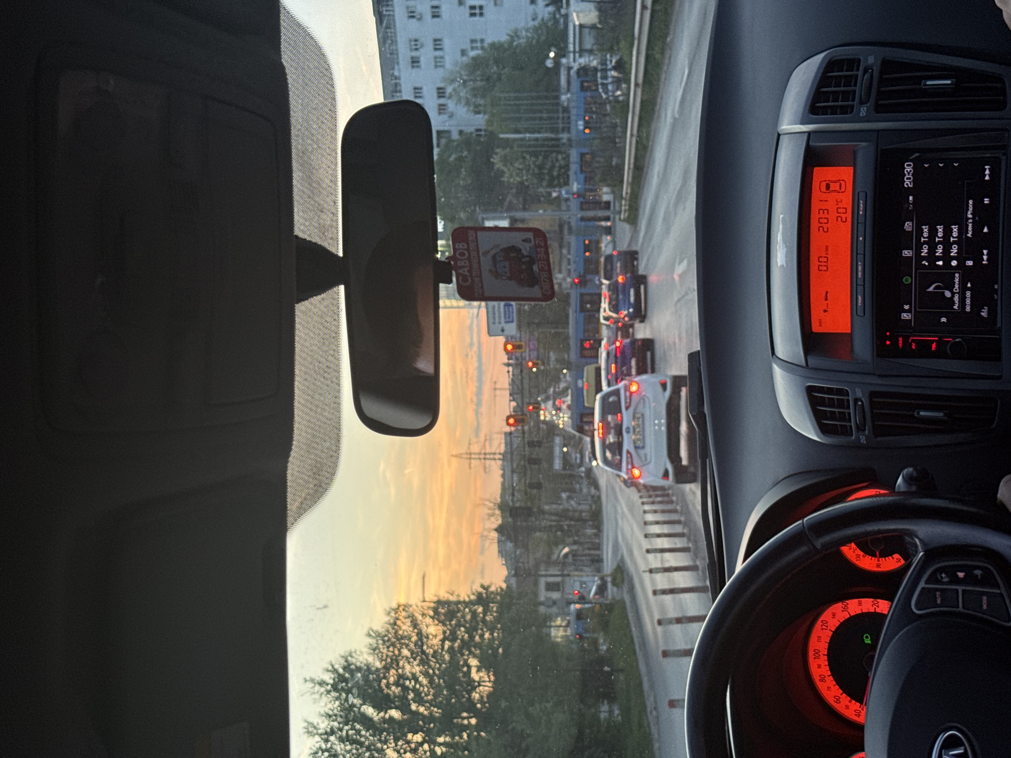 The view is from inside a car, presumably in Sofia, Bulgaria, as indicated by the "CABOB" sign with a Bulgarian phone number (0878 21 34 21) hanging from the rearview mirror. The car is stuck in traffic. The dashboard shows a temperature of 20°C and a time of 20:30. Beyond the traffic and trees, a city skyline is visible against a backdrop of a colorful sky, possibly sunset or sunrise. The steering wheel has red instrumentation lighting.