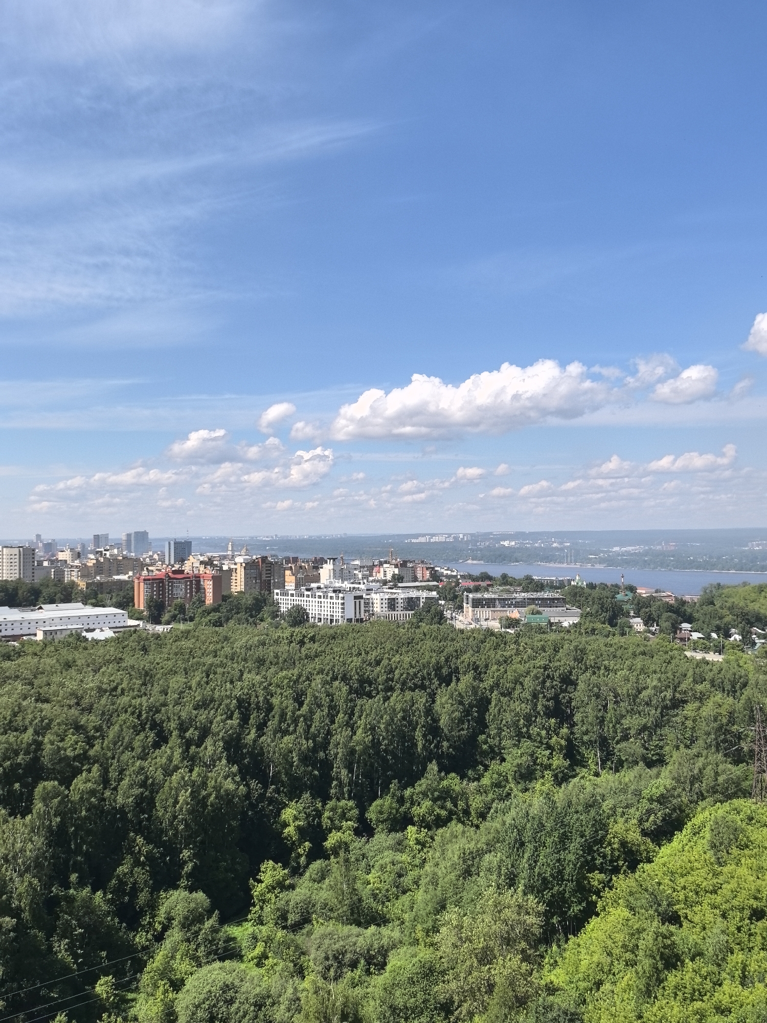 This is a wide-angle, eye-level, daytime, outdoor photograph taken from a high vantage point overlooking the city of Perm, Russia. The foreground is dominated by a dense, lush green forest covering a hillside. Beyond the forest, the mid-ground reveals a cityscape with a variety of buildings, including multi-story apartment complexes, residential buildings, and commercial structures. Some buildings are modern with glass facades, while others are older with brick exteriors in shades of orange and brown. A wide river, likely the Kama River, stretches across the horizon in the background, with hints of distant shorelines and structures visible. The sky is a vibrant blue with scattered white cumulus clouds and wispy cirrus clouds, suggesting fair weather. The overall scene depicts a harmonious coexistence of urban development and natural landscape.