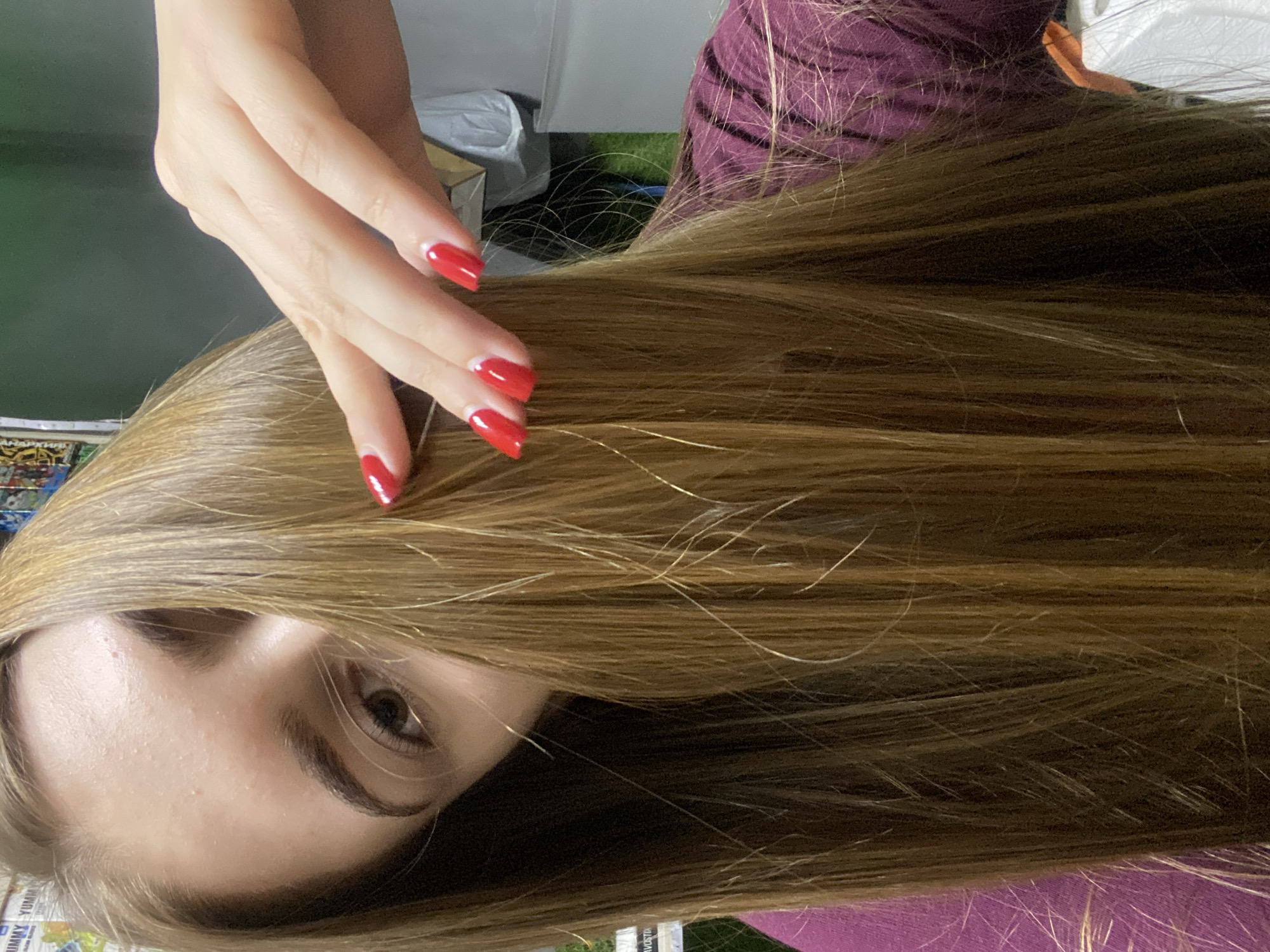 In the city of Dyakovo, Russia, a close-up image captures a young woman's face and hair. She is holding a section of her long, light brown hair with her right hand, revealing her face partially. Her fingernails are painted a vibrant red. The background is blurred, but suggests an indoor setting with hints of shelves and items on them, possibly a retail environment. The lighting is bright and natural.