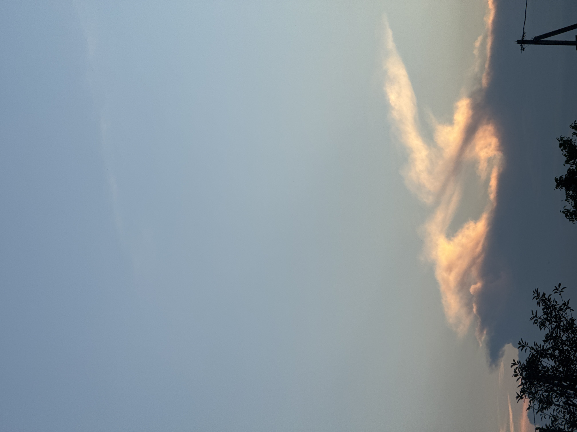 A cloudy sky with pink and orange hues is visible above a silhouetted tree line and a utility pole. The image is taken from a low-angle perspective, looking up at the sky. The clouds are dark at the bottom and lighter towards the top, with streaks of pink and orange light shining through. The sky itself is a pale blue, transitioning to a lighter hue at the top of the frame. The overall scene suggests an outdoor setting, possibly in a rural or suburban area of Бараўлянскі сельскі Савет, Belarus, during either sunrise or sunset. There are no people visible in the image.