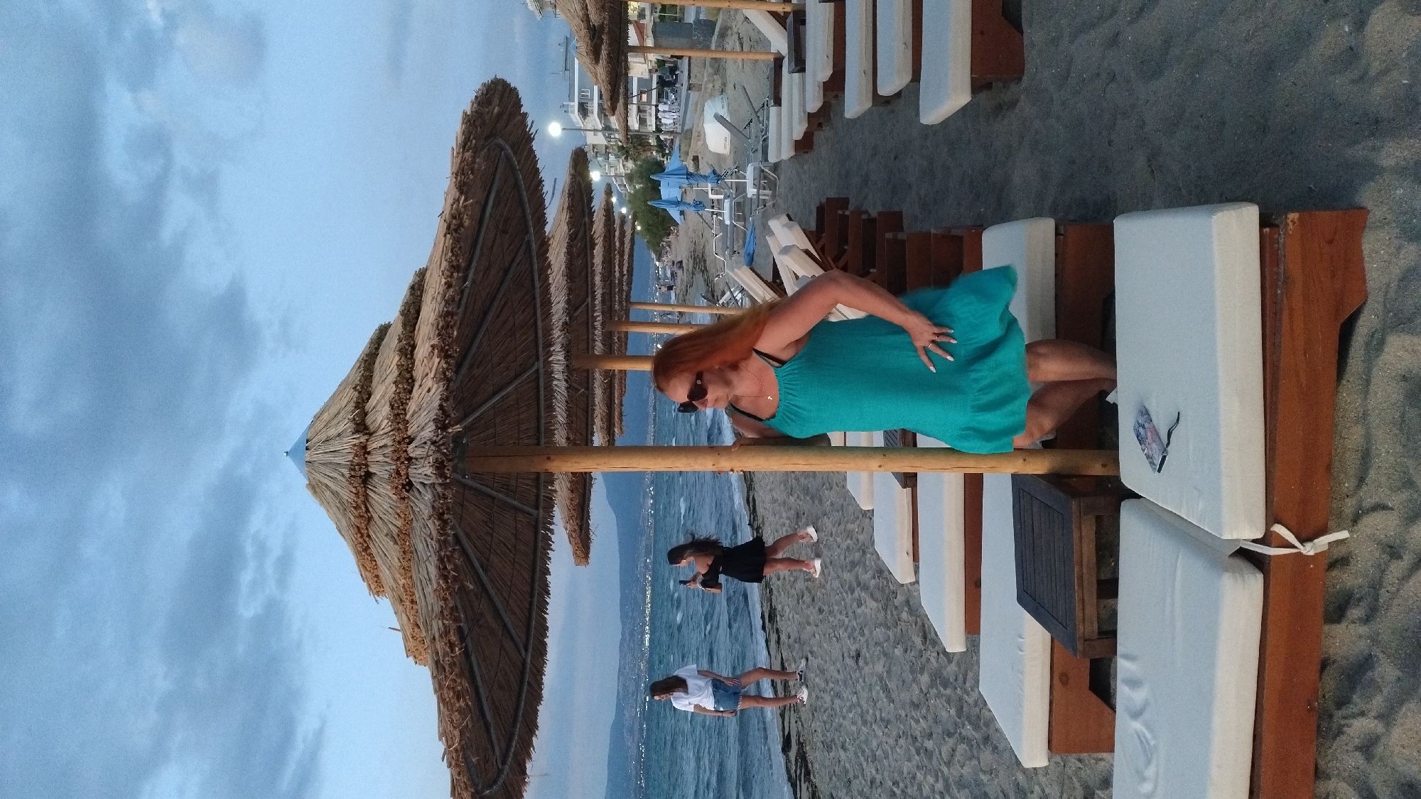 An eye-level, full shot shows a woman with long, reddish-orange hair and sunglasses standing on a sandy beach under a thatched umbrella, posing with her hand on her hip and the other on the umbrella's pole. She is wearing a teal, sleeveless dress with a ruffled hem. Behind her, several rows of empty lounge chairs with white cushions are arranged. In the background, the sea is visible with waves and distant hills. To the left, two younger women are standing near the water; one in a white t-shirt and denim shorts, the other in a black dress, holding a phone. The sky is overcast with muted blue and gray clouds. The scene appears to be a resort or public beach area in Neoi Epivates, Greece, at dusk or during an overcast day.