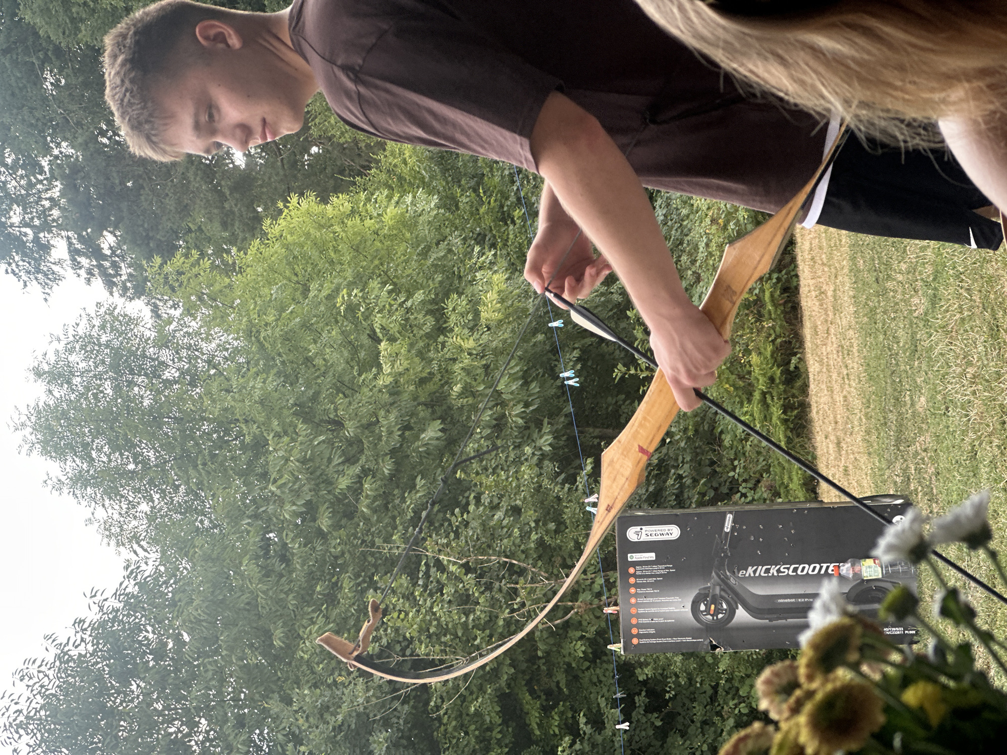 The image shows a young male, appearing to be in his late teens or early twenties, holding a wooden recurve bow and an arrow, with the arrow nocked and ready to be shot. He is wearing a dark brown t-shirt and black shorts. The setting is outdoors, with lush green trees and foliage in the background, suggesting a backyard or park environment. A cardboard box with graphics of an electric scooter is propped up as a target. There are clotheslines with clothespins strung across the scene, and some out-of-focus flowers are visible in the foreground. The lighting suggests an overcast day. The image was taken in the city of Günzburg, Germany.
