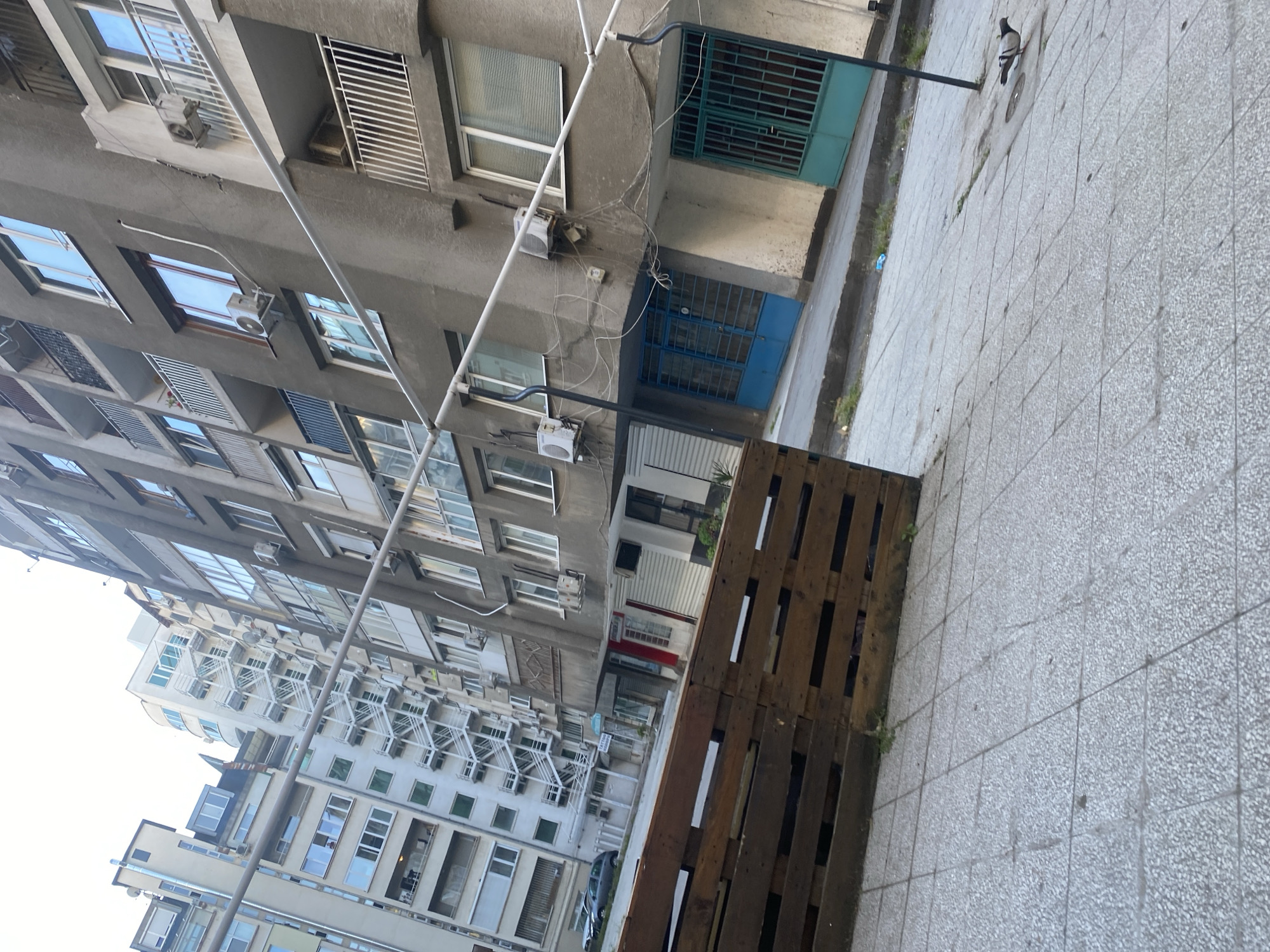 The image shows a view from a street in Sofia, Bulgaria. Several multi-story apartment buildings are visible, constructed from concrete and painted in various shades of gray and white. Many windows are equipped with air conditioning units. A wooden pallet sits on a tiled sidewalk. A pigeon is visible on the sidewalk near one of the buildings. Wires and cables are strung between the buildings. A car is parked on the street in the distance.