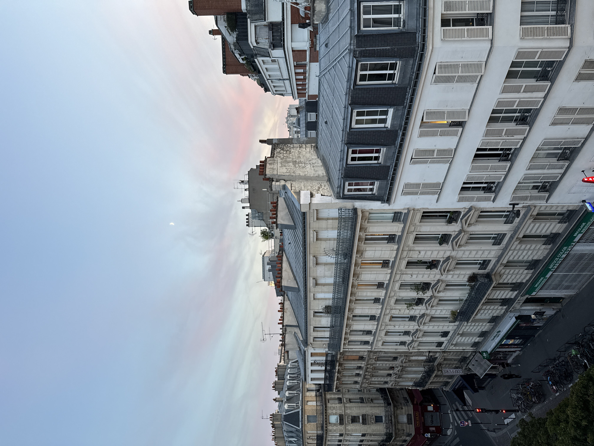 The image depicts a cityscape in Paris, France, characterized by classic Haussmannian architecture. The perspective is from an elevated viewpoint, looking down at a street lined with multi-story buildings featuring ornate facades, balconies, and mansard roofs. The sky above is a soft gradient of blue and pink, suggesting either dawn or dusk, with a sliver of the moon visible.
On the street level, there are shops and businesses, indicated by signage such as "NICOLAS," "COIFFURE," and "DROGUERIE." A traffic light is visible, showing a red signal. Bicycles are parked in racks along the sidewalk. A few pedestrians are present on the street. The buildings are densely packed, showcasing typical Parisian urban planning with balconies, numerous windows with shutters, and chimneys. The overall impression is a serene urban scene in Paris.
