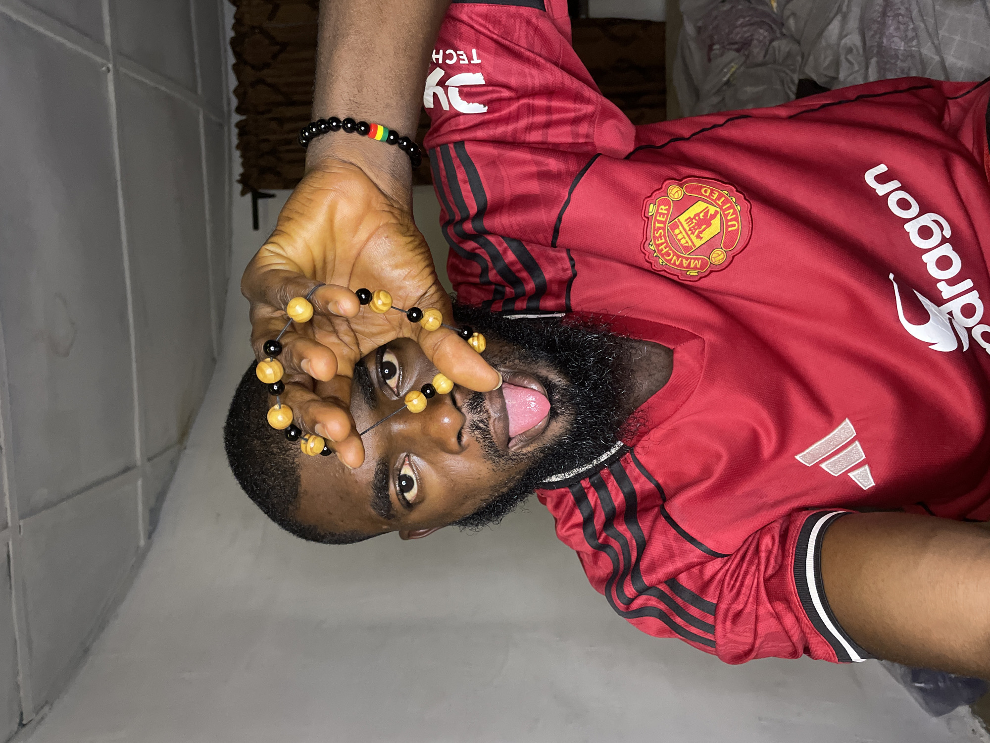 A man with dark skin, a beard, and dark hair is shown close-up from the chest up, wearing a red Manchester United jersey. He is holding a strand of yellow and black beads in his right hand, covering his left eye and part of his forehead. His tongue is sticking out. A black beaded bracelet with red, yellow, and green beads is on his right wrist. The background is dimly lit with a white ceiling and a brown curtain. The scene appears to be indoors, possibly in a bedroom, given the presence of what looks like a bed in the background. No specific location in Jos, Nigeria can be determined from the visual information.