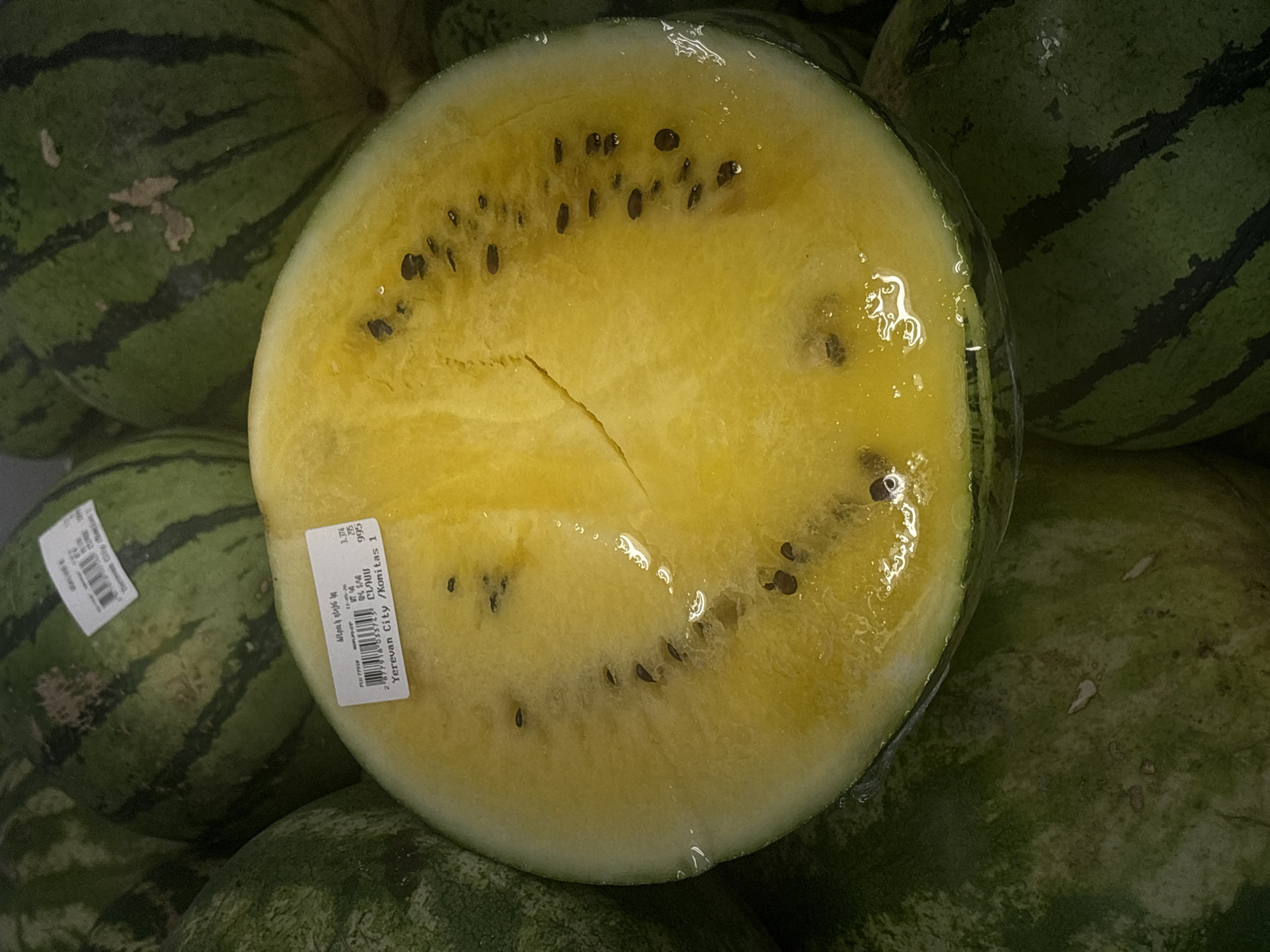The image shows a close-up of a yellow watermelon, cut in half and wrapped in plastic wrap. The watermelon is surrounded by other whole watermelons with green rinds and dark stripes. The yellow flesh of the cut watermelon contains black seeds. A price tag is visible on the cut watermelon, displaying text in Armenian and English: "ձմերուկ դեղին կա (dzmeruk deghin ka - yellow watermelon)," along with a price (995) and the location "Yerevan City / Konitas 1," which indicates the watermelon is being sold or displayed at a location within the Konitas district of Yerevan, Armenia.