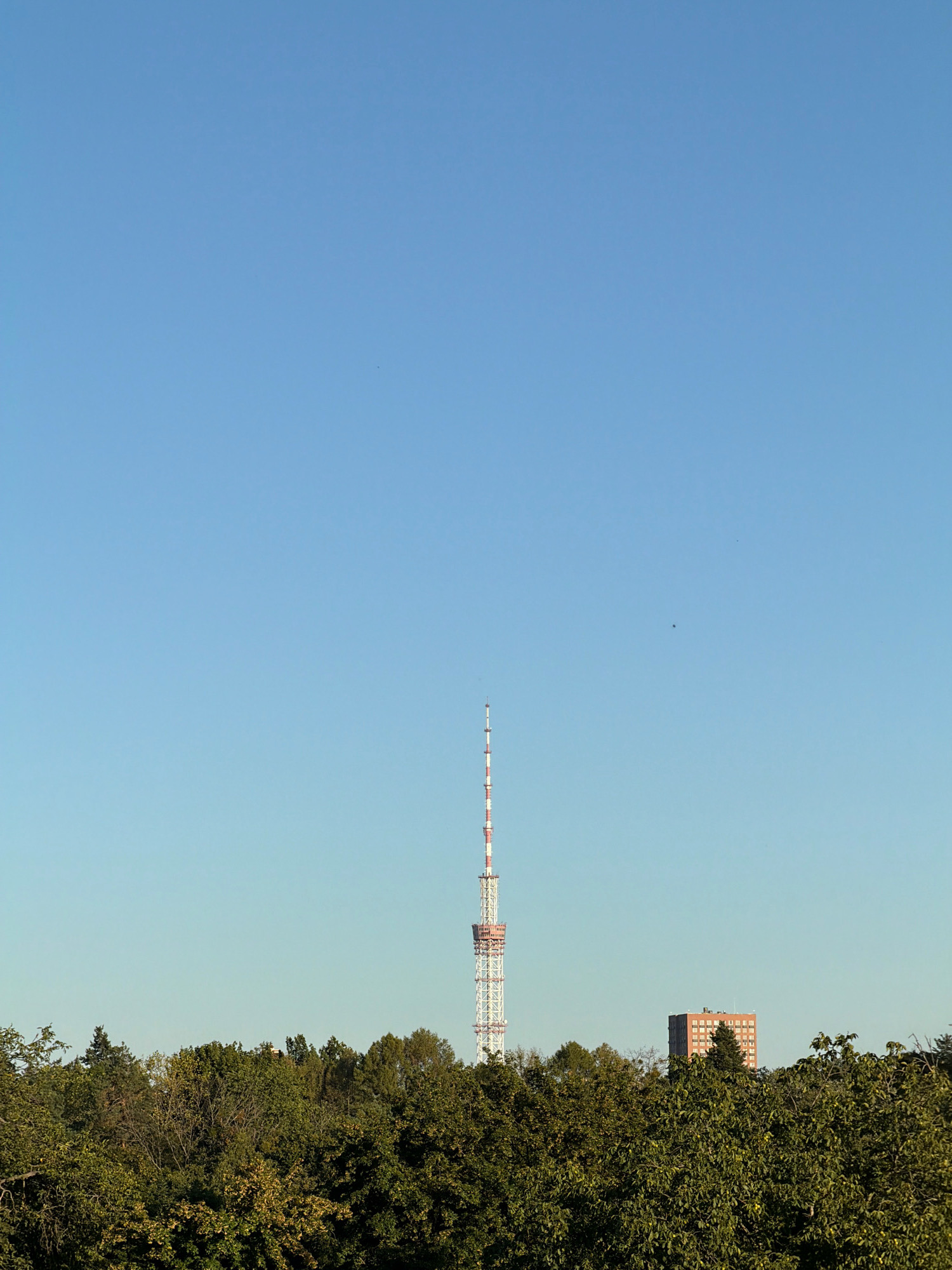 The image shows a clear blue sky above a dense canopy of green trees. A tall, slender radio tower, painted with red and white stripes, stands prominently in the center of the frame, piercing the sky. To the right of the tower, a brick-colored apartment building rises above the trees, its windows visible. The scene appears to be a view from a distance, with the trees forming a natural foreground and the tower and building silhouetted against the expansive sky, suggesting an urban or suburban setting within Kyiv, Ukraine. There are no people or discernible actions captured in the image.