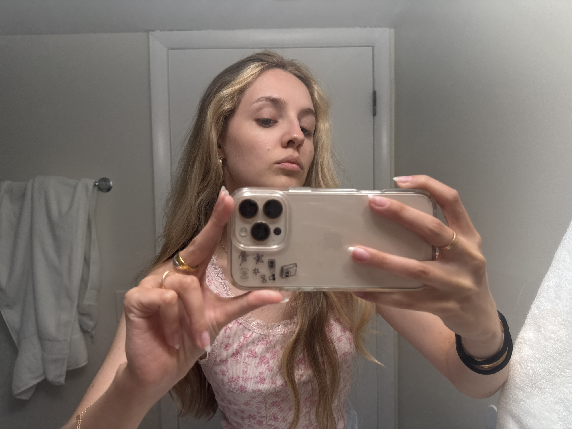 In a bathroom setting, a young woman with blonde hair takes a selfie in a mirror. She is wearing a pink floral tank top and multiple rings on her fingers. Her left hand is holding up a clear phone case with stickers on it, and her right hand is resting on her arm. A white towel is visible on a rack to her left. The background consists of a light grey wall and a white doorframe. The location is not specified in the image.
