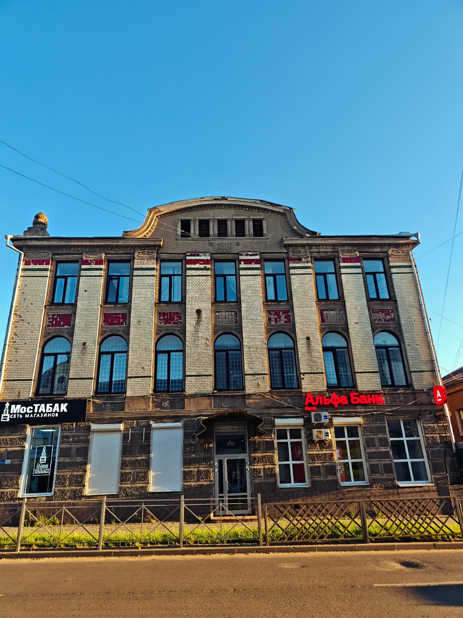 The image shows a wide, low-angle, exterior, daylight shot of a multi-story brick building. The building is located in Kimry, Russia. The facade of the building is adorned with decorative elements, including red and green stripes and carved stone details. There are numerous windows, many of which are arched. On the first floor, there are storefronts. A sign with Cyrillic text reads "МосТАБАК" and another sign with red Cyrillic text reads "Альфа Банк". A metal fence runs along the front of the building, with a strip of green grass and a sidewalk behind it. A paved road runs in front of the sidewalk. The sky is clear and blue. Electrical wires are visible against the sky.