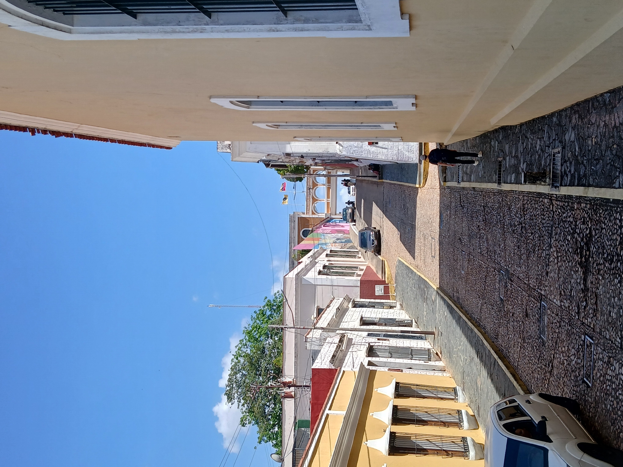 A street view in Ciudad Bolivar, Venezuela, shows a narrow cobblestone street flanked by buildings with yellow and white facades. The sky is clear and blue. Towards the end of the street, an arched structure is visible with flags. A man walks on the right sidewalk while a car drives on the street. A white car is parked on the right side of the frame. Various windows and architectural details are evident on the buildings.