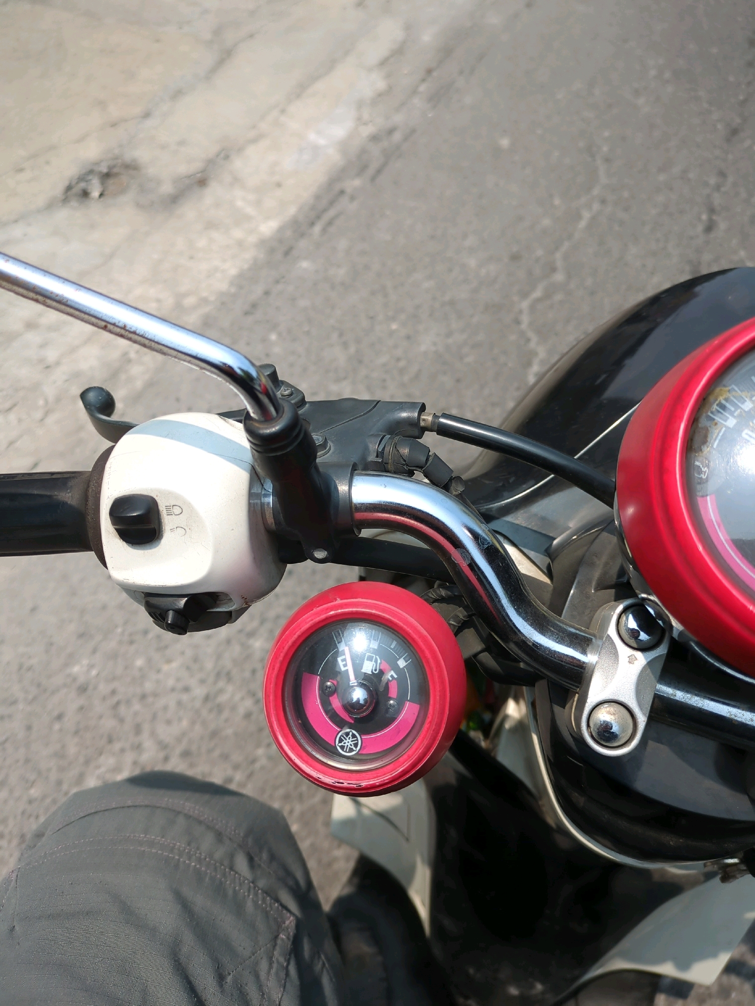 The image is a close-up, first-person perspective shot of a motorcycle's handlebars and a portion of the rider's leg. The motorcycle's instrument panel, featuring a fuel gauge with a needle indicating a nearly full tank, is clearly visible. To the left of the fuel gauge is a white control housing with a black toggle switch for lights. A chrome handlebar extends across the frame, and parts of the motorcycle's black bodywork are visible. The background is an asphalt road, suggesting an urban environment, consistent with the general setting of Surakarta, Indonesia. The lighting indicates it is daytime, likely outdoors. No other individuals are present in the frame.