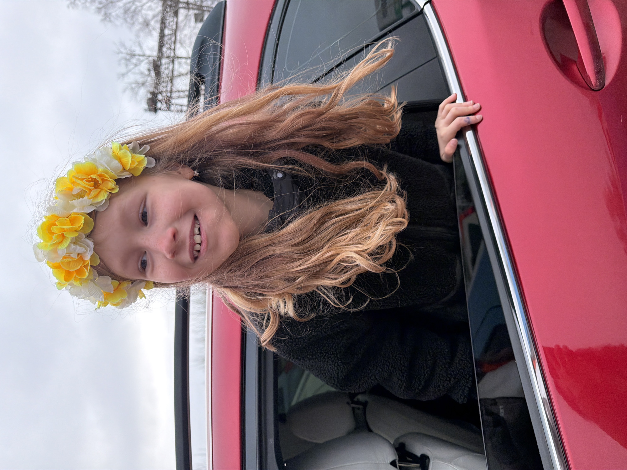 A young girl with long, wavy blonde and light brown hair is captured leaning out of the passenger window of a vibrant red car. She is smiling broadly and happily, with her hands gripping the top edge of the car's door frame. Her head is adorned with a festive flower crown composed of white and yellow artificial flowers. She is wearing a black, fuzzy jacket or coat.
The car, which appears to be a sedan or SUV, has shiny red paint and a dark-tinted window. Partially visible through the car window are light-colored, possibly white or grey, car seats. The background features an overcast, light grey sky, indicating a cloudy day. Bare trees stand in the distance, suggesting a cooler season such as late autumn, winter, or early spring. A tall, skeletal structure, possibly a power line tower or crane, can be faintly seen through the trees in the upper left. The overall mood conveyed by the girl's expression is one of joy and excitement. This scene takes place in Chaikovsky, Russia. There is no visible text in the image.