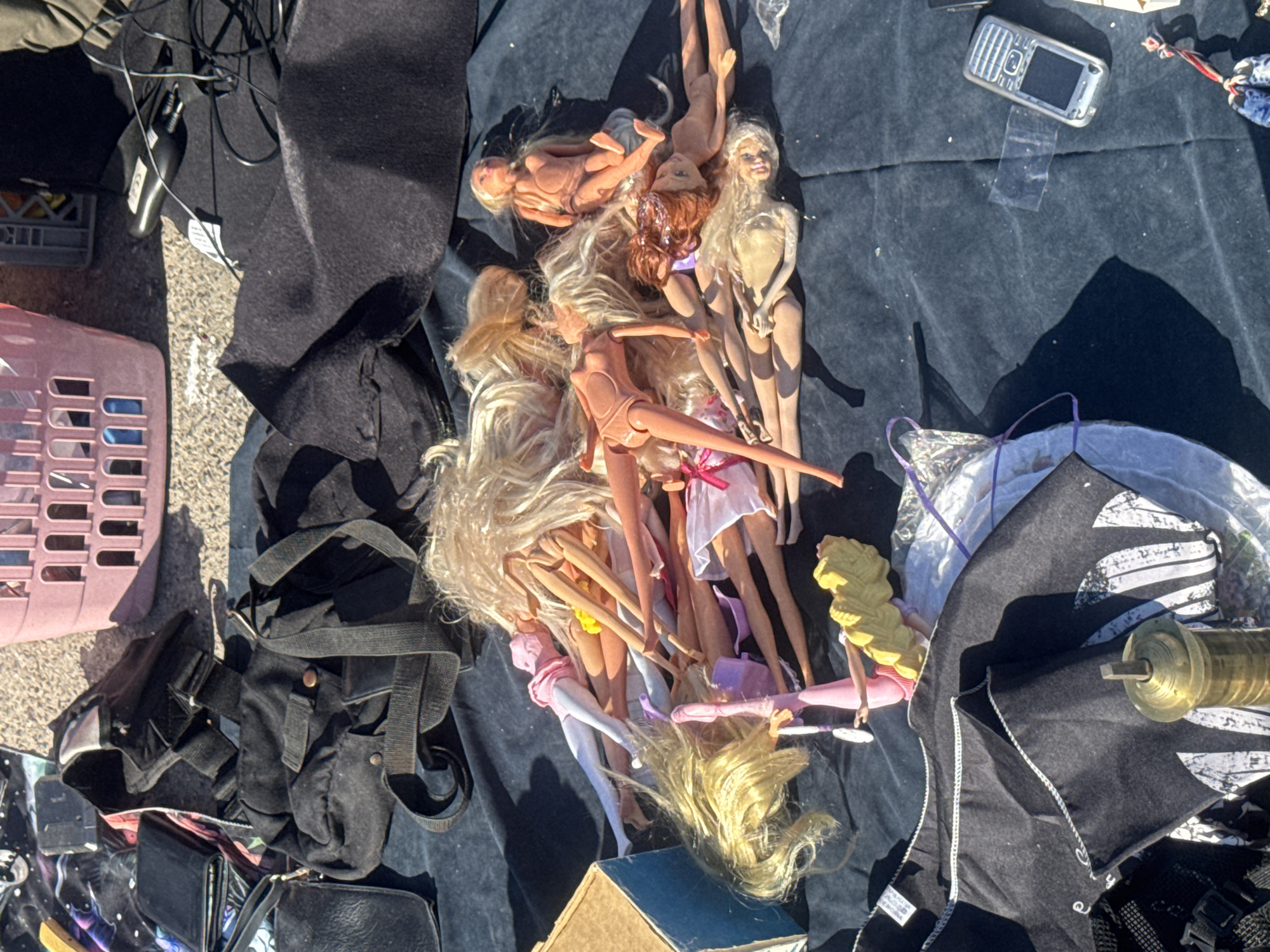 A pile of Barbie dolls is spread out on a dark blue fabric surface. The dolls have blonde, brown, and reddish hair and are wearing various clothing items including pink and white dresses. Some of the dolls are partially undressed. In the background, there is a pink plastic basket, a black bag, and other assorted items. The scene appears to be outdoors, possibly at a market or flea market, given the presence of multiple items for sale. The lighting suggests it is daytime, with shadows cast on the fabric.
