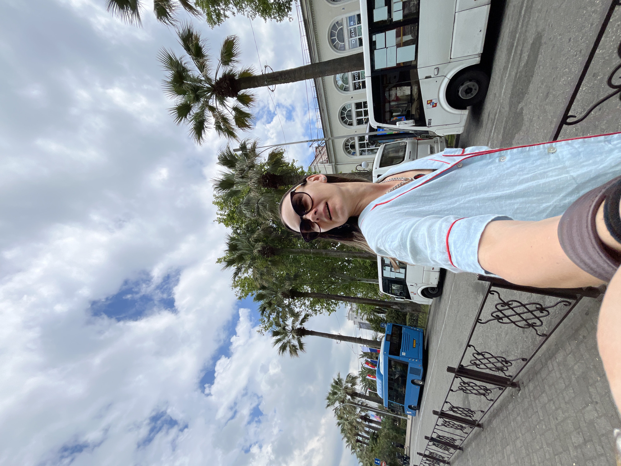 In Sochi, Russia, a woman with sunglasses and a light blue pajama-like outfit stands on a sidewalk. Palm trees line the street, and a blue bus is parked in the background. The woman is looking away from the camera, with a slight smile on her face. The sky is cloudy with patches of blue visible. A decorative metal fence runs along the sidewalk.