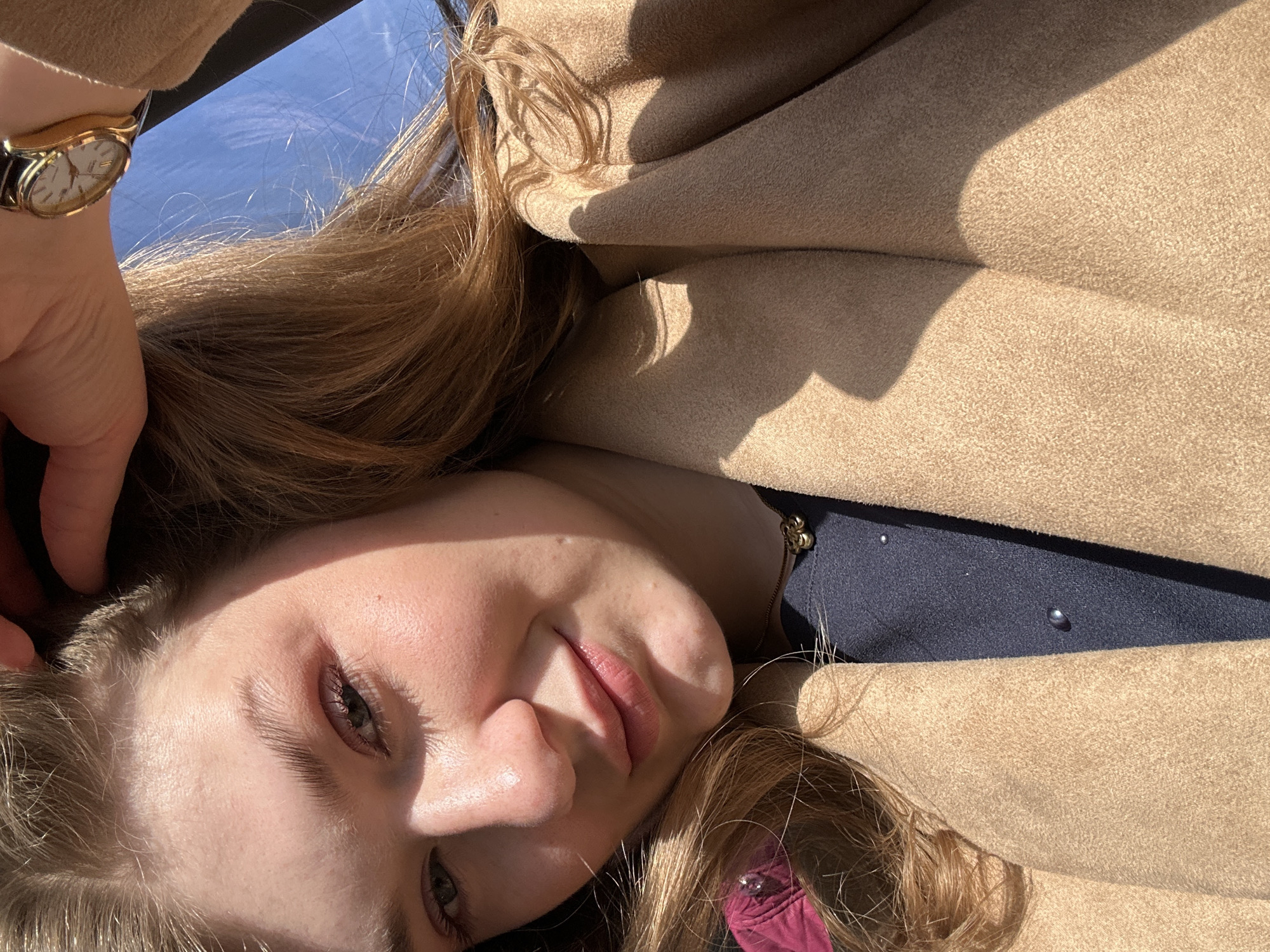 A young woman with fair skin and long, light brown hair is positioned at an oblique angle in the frame, her face close to the camera. She is wearing a dark blue top or dress with pearl-like embellishments, underneath a tan, wool-like coat or jacket. A gold-colored wristwatch is visible on her left wrist.
The location is the city of Kazan, Russia, although no specific landmarks or architectural details are visible to confirm this. The background shows a bright blue sky suggesting the photo was taken outdoors on a sunny day. The woman is possibly in a vehicle or in transit, given the angle and composition of the shot.