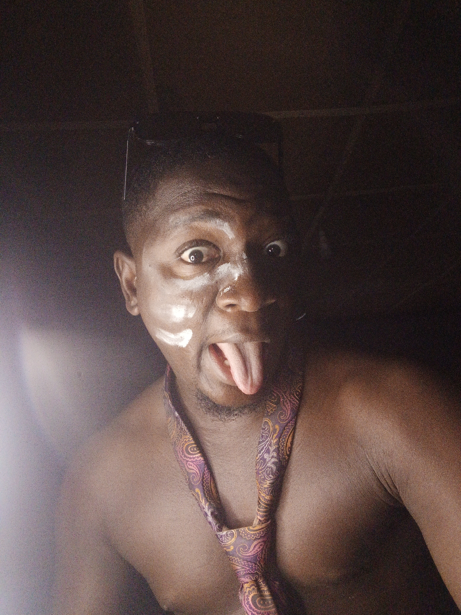A poorly lit close-up photo shows a dark-skinned individual with white markings on their face, eyes wide, and tongue sticking out. They are wearing a patterned tie loosely around their neck and appear to be shirtless. Sunglasses are visible on top of their head. The image appears to be taken indoors. There is no information available to determine the location in Akwanga, Nigeria.
