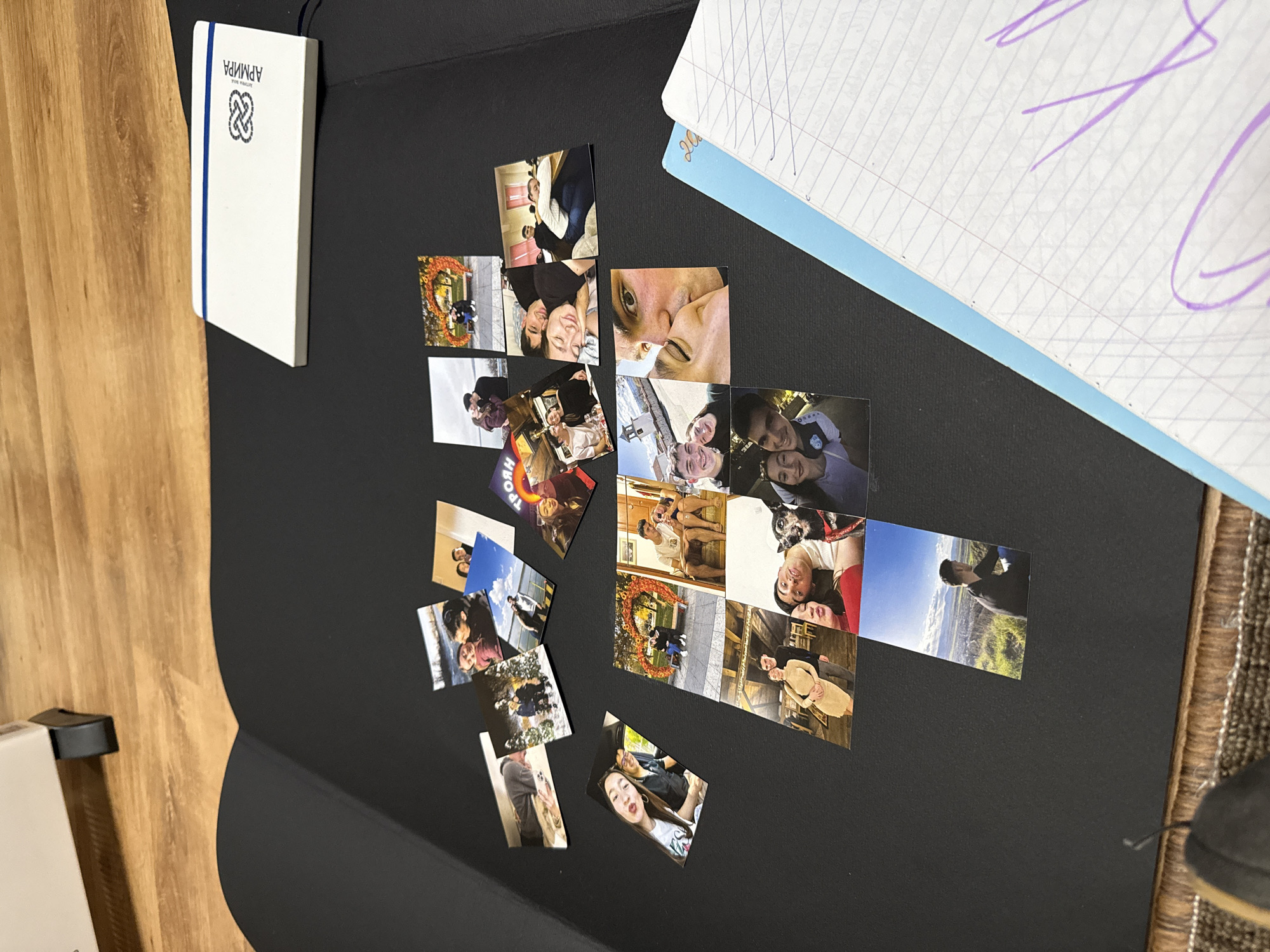 The image shows a collection of photographs arranged on a black surface, likely a table or board. The photos appear to depict a couple in various settings and poses. To the left, there is a white notebook with a blue spine and a logo in the upper corner. A second notebook with purple markings is to the right of the photos. The background consists of wooden flooring, and a patterned rug. The location is likely an indoor setting in Sofia, Bulgaria, based on the Cyrillic writing on the white notebook.