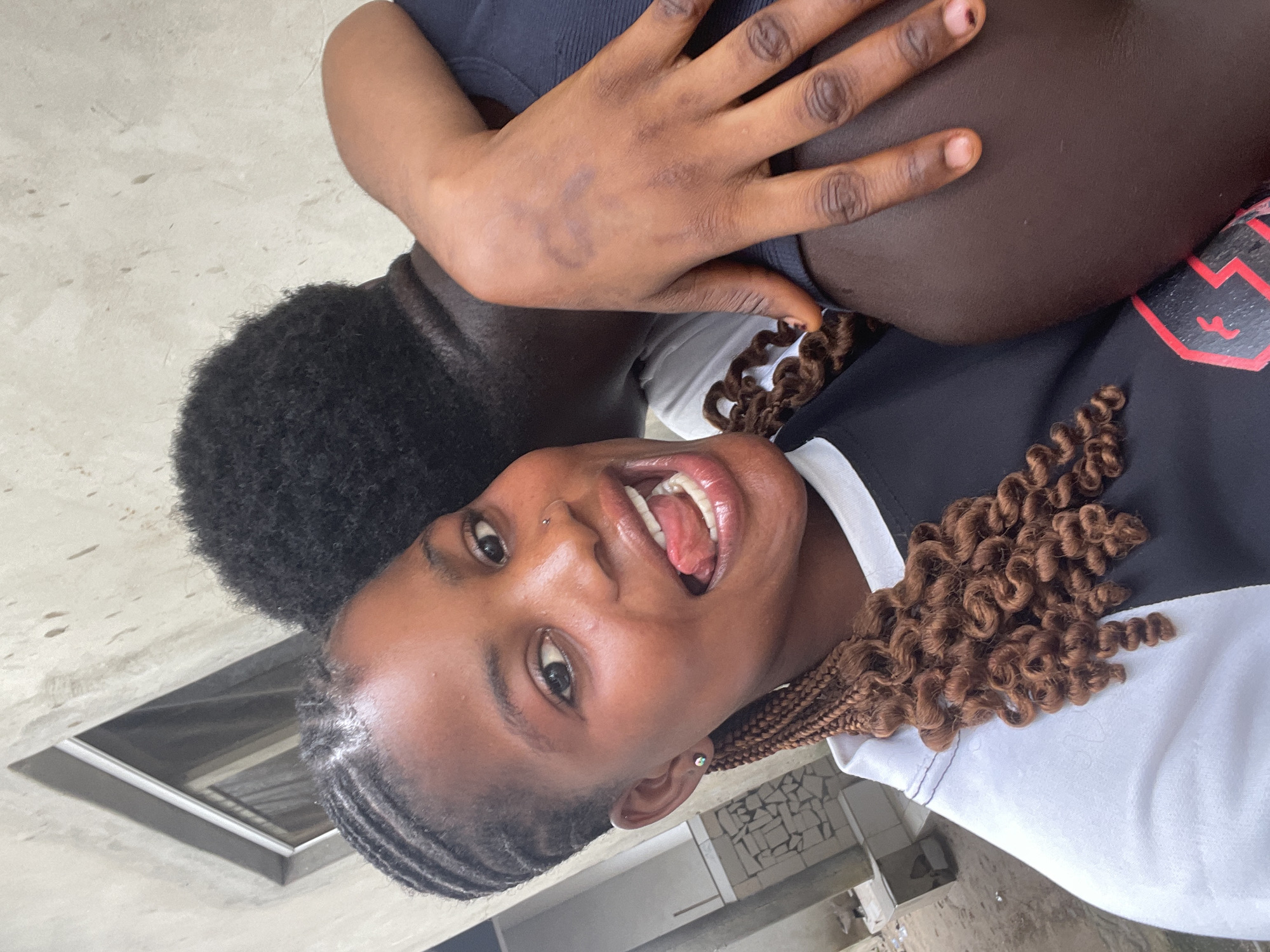 A close-up, portrait-oriented image taken in Badagry, Nigeria, features two individuals in close proximity, likely during the daytime. The photograph is slightly rotated clockwise.
The primary subject, a person with dark skin, is in the foreground, looking directly at the camera with a playful expression. Their mouth is open, revealing white teeth, and their tongue is slightly extended. They have dark braided hair, with cornrows at the front and voluminous, curly brown extensions on the side/back. Notable details include a small silver stud nose piercing and a small earring. This person is wearing a two-toned top, black on top and white below, with a red graphic design that appears to be a stylized number or symbol on the black section.
Behind and to the side of the first individual, a second person, also with dark skin, is partially visible. Their left arm is draped over the shoulder of the foreground subject, with their hand resting on the upper arm. This second person has dark, curly, Afro-textured hair and is wearing a dark-colored, possibly ribbed, top. A faint, dark mark, somewhat circular in shape, is visible on the back of their hand.
The background consists of a plain, light-colored surface, possibly a concrete wall or floor, with a dark rectangular opening visible in the lower left quadrant, which might be a window or doorway. The overall emotion conveyed by the primary subject's expression is one of playfulness or joy. No legible text is visible in the image.
