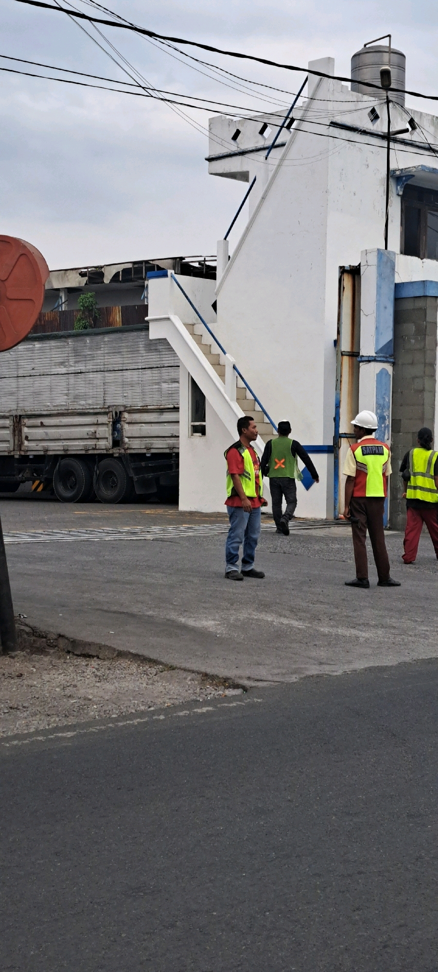 The image depicts an outdoor scene in Pandaan, Indonesia, likely near an industrial or commercial area. In the foreground, a grey asphalt road with some cracks and gravel is visible. Several individuals are present, all wearing high-visibility vests over their clothing.
To the left, a large, silver truck with a grey cargo bed is parked. A red circular object is partially visible on the far left edge of the frame, positioned above the truck.
The background features a white building with blue accents and an external staircase. Overhead, a network of black electrical wires crisscrosses the sky, which is overcast.
The individuals are the primary focus of the scene. From left to right:
1. A man in a red shirt and a yellow high-visibility vest stands facing left, looking forward. He is wearing blue jeans and dark shoes.
2. A person wearing a dark long-sleeved shirt and a green high-visibility vest with an orange "X" design on the back is walking away from the camera. They are holding a blue folder or clipboard.
3. A person wearing a light-colored shirt, a dark vest over it, a white hard hat, and a yellow high-visibility vest with the word "SATPAM" printed on it is facing the camera. They are wearing brown pants and black shoes.
4. Another person wearing a dark shirt and a yellow high-visibility vest is visible to the far right, with their back to the camera. They are wearing red pants and black shoes.
The overall impression is that of workers or security personnel engaged in some activity on a street adjacent to a building and a parked truck.