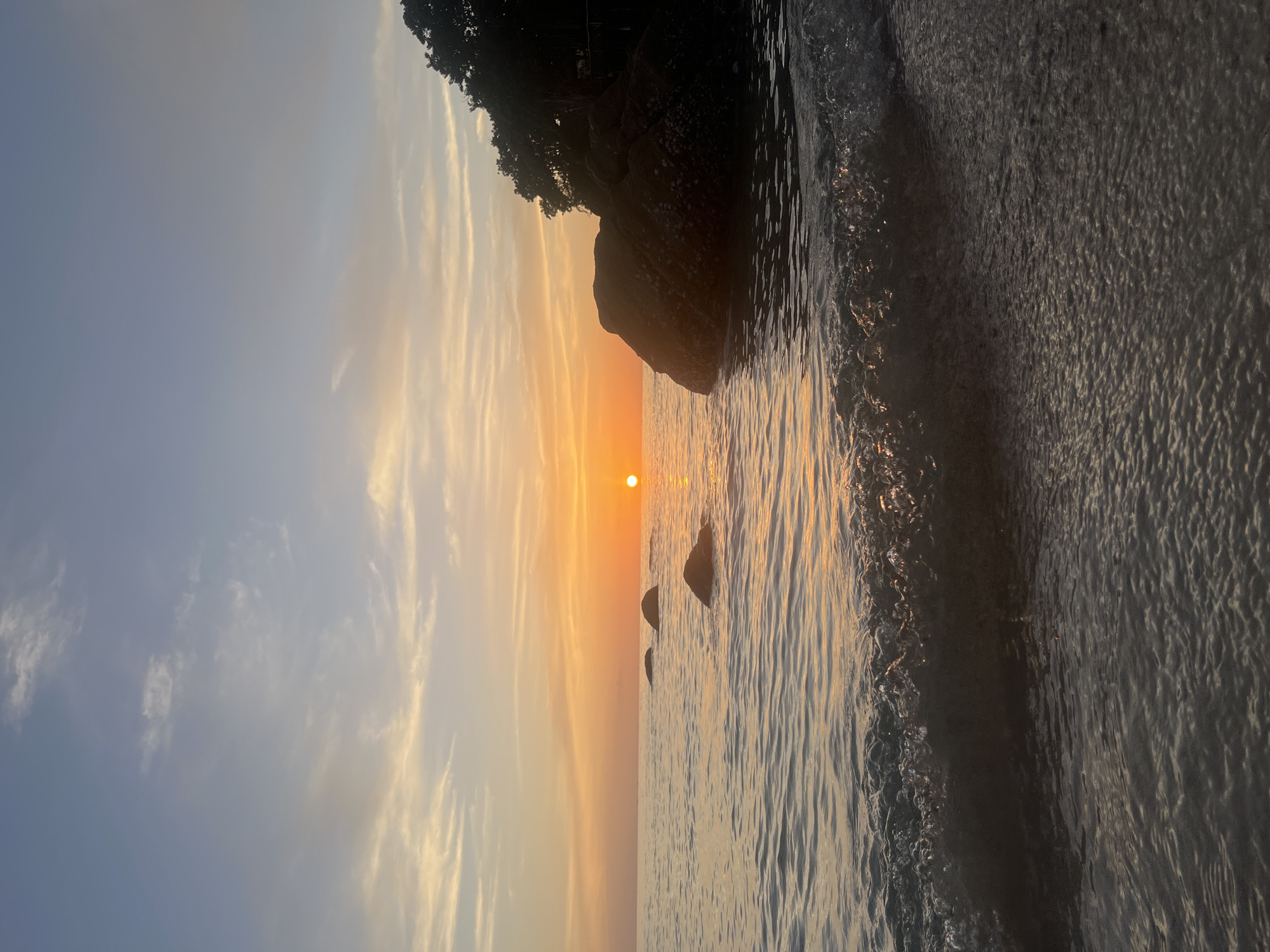 A serene sunset paints the sky with hues of orange and blue over the ocean in Ko Pha-ngan District, Thailand. The sun, a bright orange orb, is low on the horizon, casting a warm glow on the water. Gentle waves lap at the sandy shore, with a small wave cresting in the foreground, its foam catching the light. Several dark, round rocks protrude from the calm sea, appearing like sentinels against the vast expanse of water. To the right, a large, dark rock formation juts out of the water, its surface dotted with barnacles. Behind the rock, a dense silhouette of lush green trees lines the coastline, their foliage forming a dark, shadowy mass against the vibrant sky. The sky itself is a canvas of soft, wispy clouds, streaks of white and pale yellow scattered across the gradient of blues and oranges. The overall scene evokes a sense of peace and tranquility, a perfect end to a day on this Thai island.