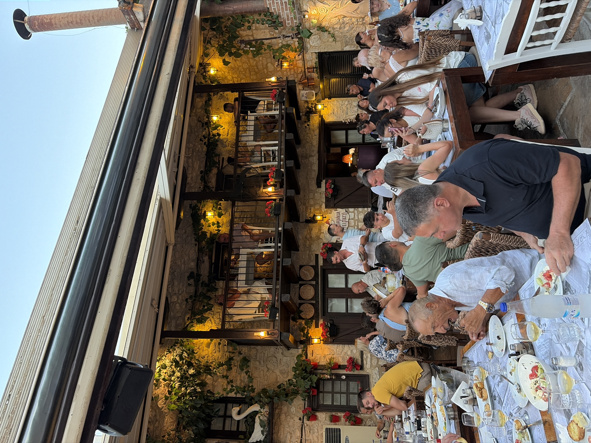 The image depicts an outdoor dining scene at a restaurant in the Kassandra Municipal Unit, Greece. Multiple tables are set with food and drinks, and a diverse group of people are seated and interacting. The architecture suggests a traditional Greek style, with stone walls, wooden balconies adorned with plants and flowers, and outdoor lighting. In the foreground, an elderly man is eating, while other patrons are engaged in conversation and enjoying their meals. The atmosphere appears casual and convivial.