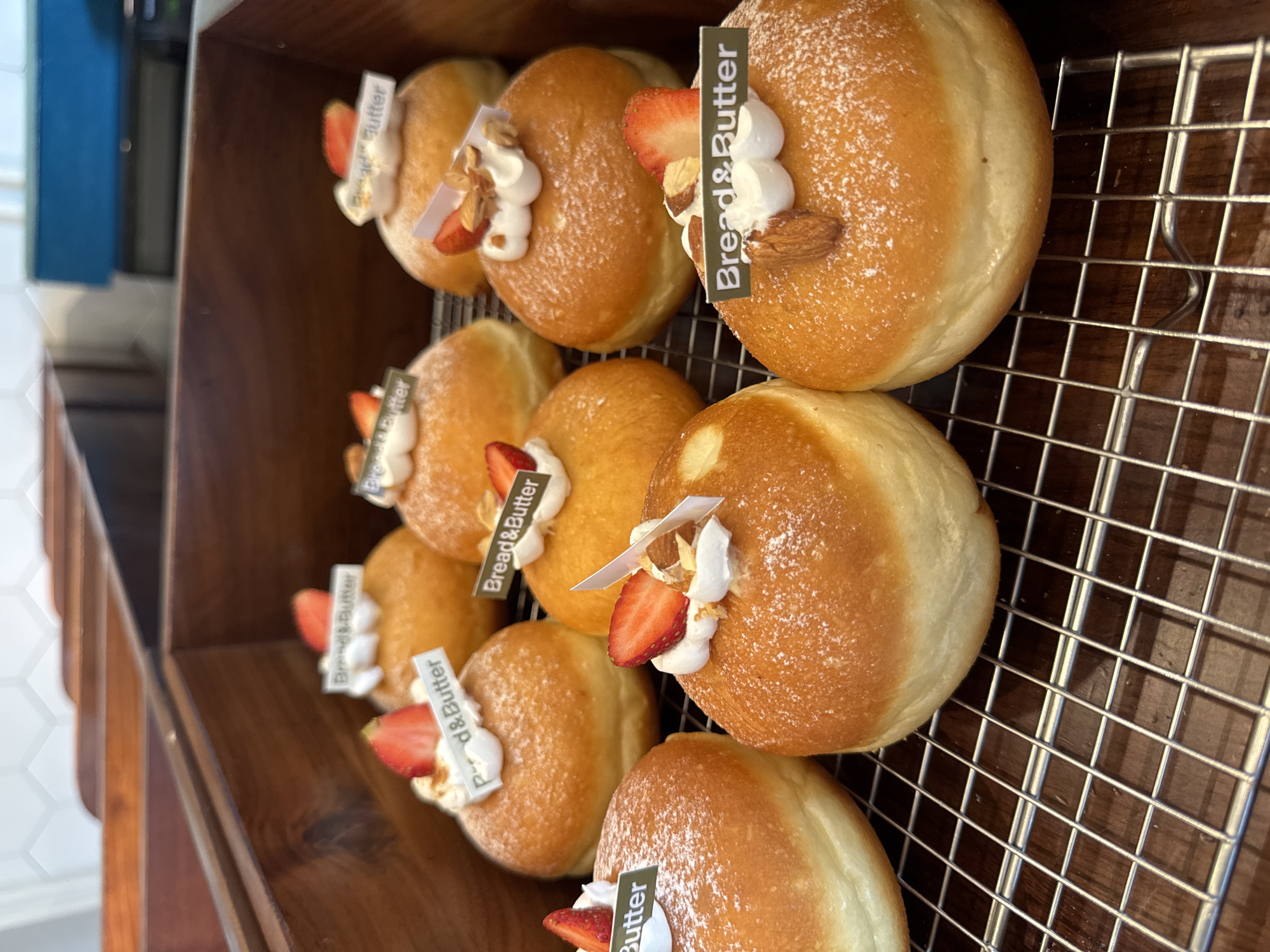 A close-up, slightly high-angle shot shows a display of several donuts or cream-filled pastries. Each pastry is topped with a dollop of white cream, a slice of strawberry, and is adorned with a small, rectangular tag labeled "Bread & Butter". Some pastries also include a whole almond. The pastries are arranged in rows on a wire rack, which is placed inside a dark wooden display case. The focus is sharp, highlighting the textures of the sugary pastry, the creamy topping, and the fresh fruit. The color palette is warm, with golden browns of the pastries contrasting with the bright red of the strawberries and the white of the cream. The image evokes a sense of indulgence and craftsmanship. The pastries appear to be on display for sale, perhaps in a bakery or cafe. A plausible location is a cafe or bakery in Đà Nẵng, Vietnam, based on the prevalence of similar pastries in Southeast Asian cafes.