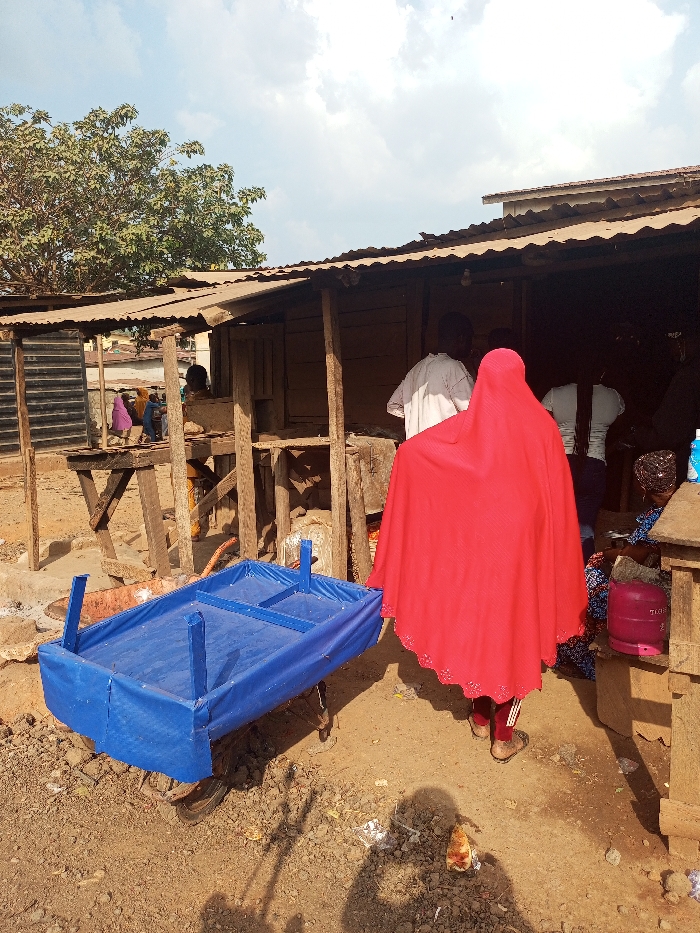 This image, taken in Aramoko, Nigeria, depicts an outdoor scene characterized by informal commercial activity under a bright, partly cloudy sky. In the foreground, a blue, fabric-covered, two-wheeled handcart or vending trolley is positioned on unpaved dirt ground, facing left. The ground shows scattered debris, including a discarded piece of food and white paper.
In the mid-ground, a person wearing a long, red, floor-length robe or hijab with a scalloped edge and dark leggings with sandals is seen from behind, positioned slightly right of center. This individual faces towards a collection of makeshift wooden stalls or shelters with corrugated metal roofs. Behind and to the right of this person, another individual in a white, long-sleeved top is partially visible. Further right, a person is seated on a low stool or bench, next to a pink gas cylinder.
The background is dominated by a series of rudimentary wooden structures with corrugated metal roofs, supported by rough wooden posts. Within these structures and further back, several other individuals are visible, including figures in purple and blue garments on the far left. A large, leafy green tree is prominent on the left side, extending above the structures. Distant rooftops are visible under the horizon line. The overall environment suggests a bustling, unpaved commercial or community area.