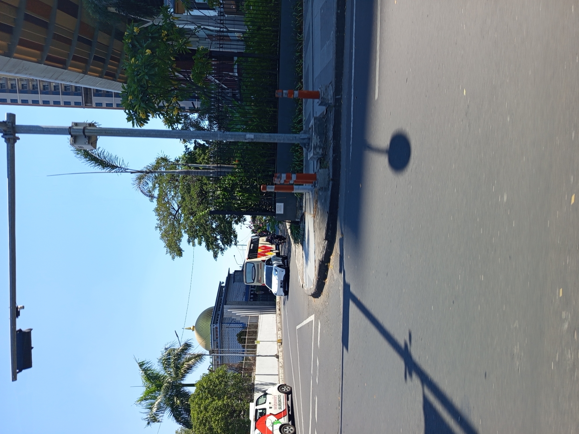 The image captures a street scene in Surabaya, Indonesia, on a clear, sunny day. In the foreground, a grey asphalt road with white lane markings is visible, with the shadow of a traffic light pole cast across it. The road appears to be an intersection, with a curved curb on the right side.
To the left, a white van with red and green decals, displaying the text "Wirawiri SUROBOYO," is parked or moving. Behind the van and across the street is a building with a golden dome, characteristic of a mosque. The building has intricate blue and white tilework on its facade. Palm trees and other lush green foliage surround the mosque.
Further down the road, a white car, possibly an electric vehicle due to its sleek design, is turning. A large bus with a white and orange livery is also present on the road, as is a motorcyclist. In the background, a tall, modern building with multiple floors rises into the clear blue sky, juxtaposed with the more traditional architecture of the mosque. A black metal fence with dense green vegetation behind it lines the right side of the scene. Orange traffic cones are placed along the curb on the right, indicating potential road work or traffic management. The overall impression is a bustling urban environment where modern infrastructure meets cultural landmarks.