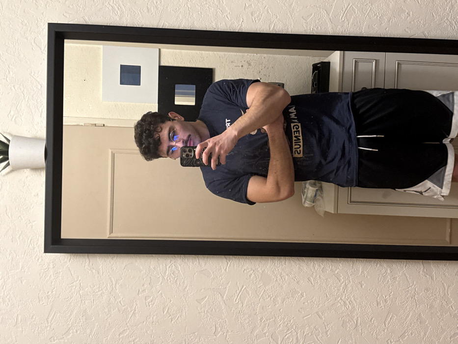 This image depicts a mirror selfie taken indoors in Rennes, France. A male individual, appearing to be an adult, is visible from the waist up, reflected in a large, black-framed mirror. He has dark, curly hair, a mustache, and is wearing round spectacles that reflect blue light. He is dressed in a dark blue short-sleeved t-shirt with reversed text (including "GENIUS") and black athletic shorts featuring white and grey panels with a visible Jordan logo. His arms are crossed, and he is holding a dark-colored smartphone with a textured case in his right hand, positioned to capture his reflection.
The environment reflected in the mirror includes a light-colored closed door to the left of the individual. Below his waist, a white vanity or chest of drawers is visible, with a black tray and other indistinct items on its surface. Above the vanity, two pieces of wall art are displayed: a white square frame containing a dark blue square, and below it, a black square frame with multiple light blue vertical stripes. A small white potted succulent rests on the top edge of the mirror frame. The wall surrounding the mirror is textured with a light, off-white finish.
