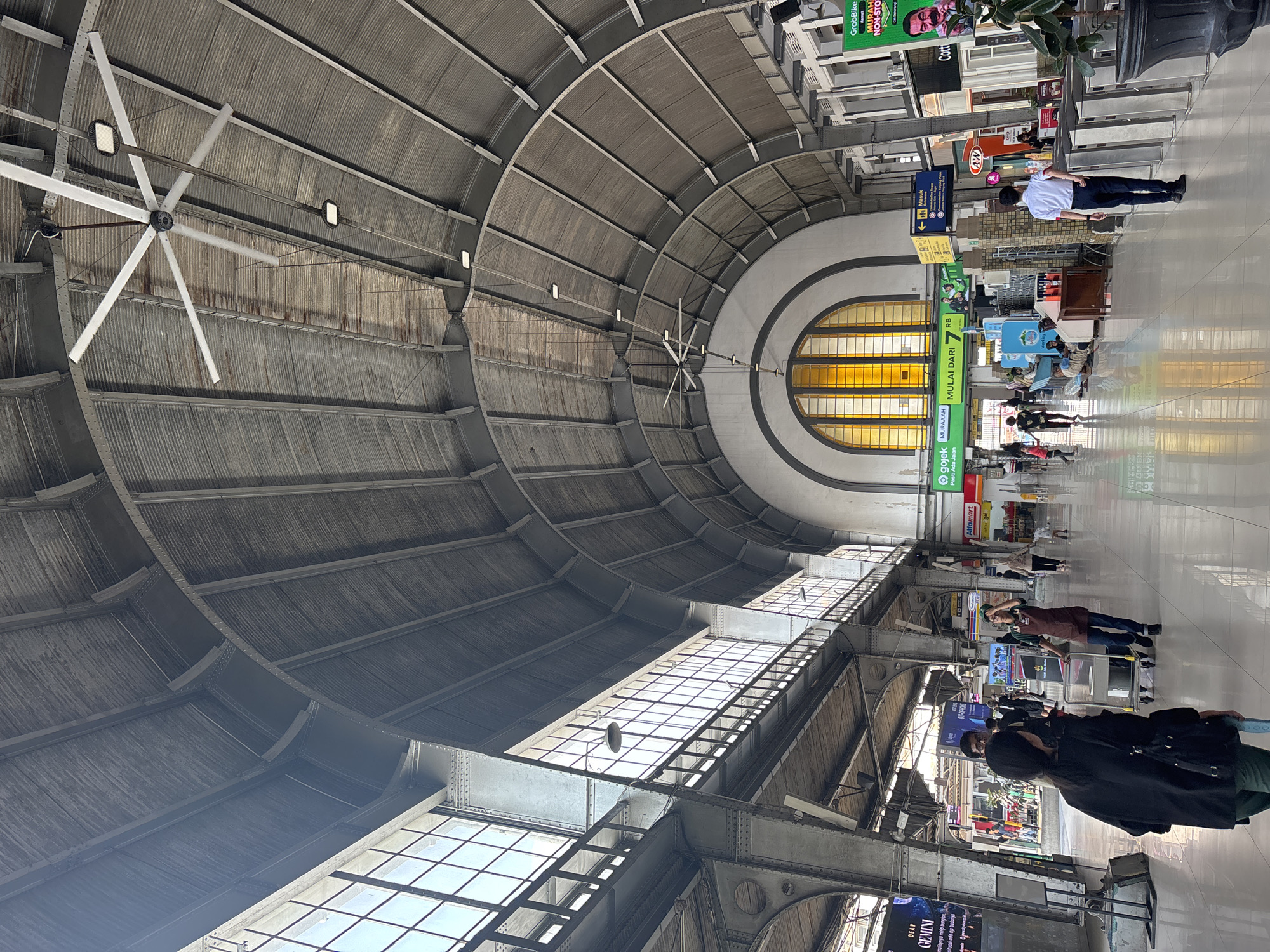 The image depicts the interior of a large, cavernous building, likely a train station, with a high, arched, corrugated metal ceiling supported by exposed steel beams. Large windows with a grid pattern are visible along the left side, letting in natural light. Several large industrial ceiling fans are suspended from the ceiling. The floor is polished and reflective, showing the architectural details of the ceiling.
In the foreground, a woman with dark hair, wearing a black jacket and carrying a backpack, is walking away from the camera. To her right, a man with glasses and a dark shirt is walking towards the camera. Further into the scene, there are various people, some waiting, some walking, and some pushing carts. Signs for services like "GoJek" and "Alfamart" are visible, as well as advertisements. In the background, a large arched window with yellow panes is a prominent feature. The overall impression is of a busy public space with historical architecture. This appears to be the interior of Gambir Station, a major railway station located in Gambir, Gambir, in the city of West Jakarta, Indonesia.