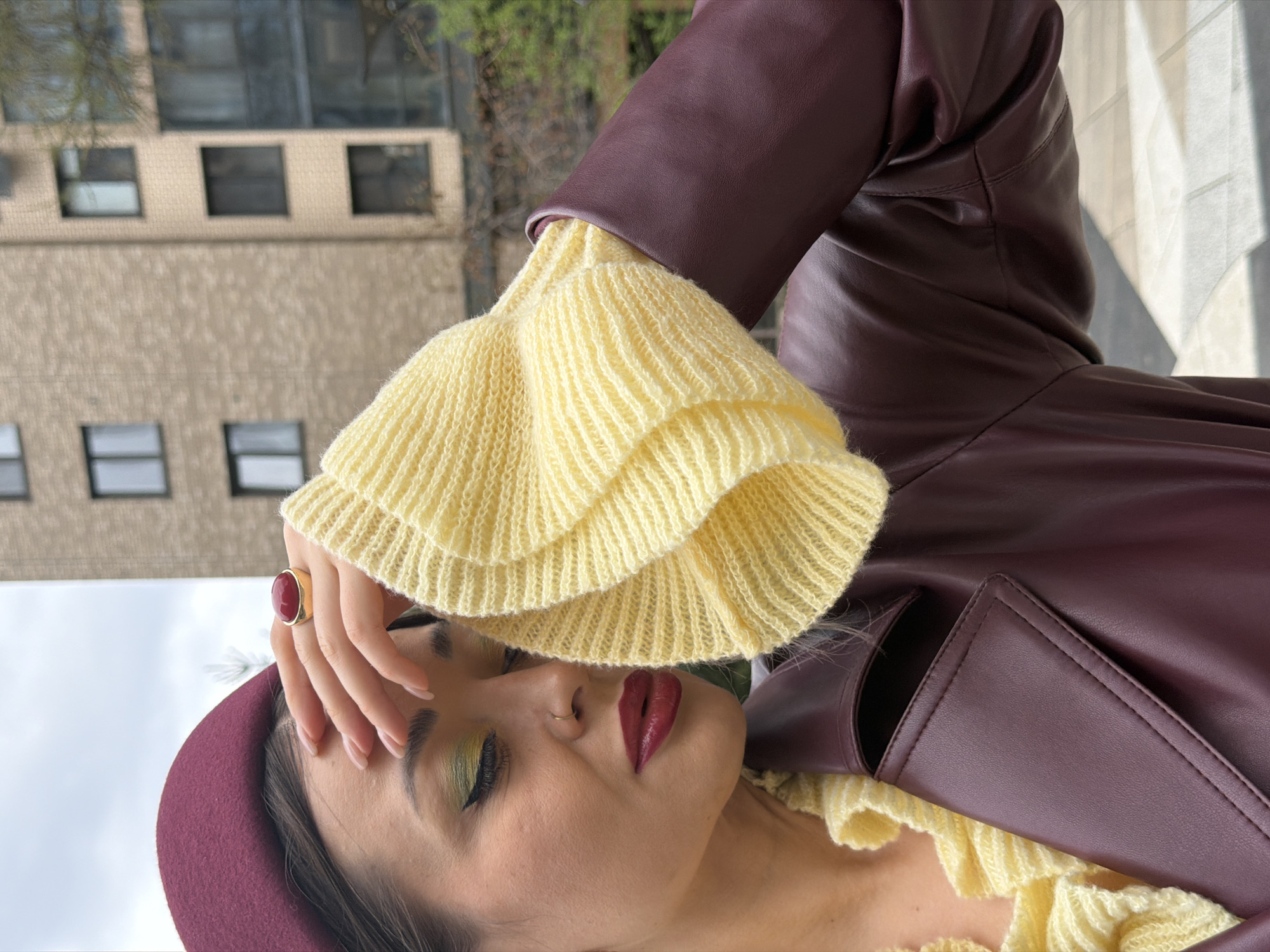A woman is depicted in an outdoor shot, wearing a burgundy beret, a burgundy leather jacket, and a yellow ruffled knit top. Her face is partly obscured by her hand, which is adorned with a ring featuring a large red stone. She has dark hair, green eye shadow, and burgundy lipstick. The background includes a building with rectangular windows and a sky with what appears to be clouds. The photo seems to be taken outdoors during daylight hours. The location, Krasnoyarsk, Russia, is not discernible from the image alone.