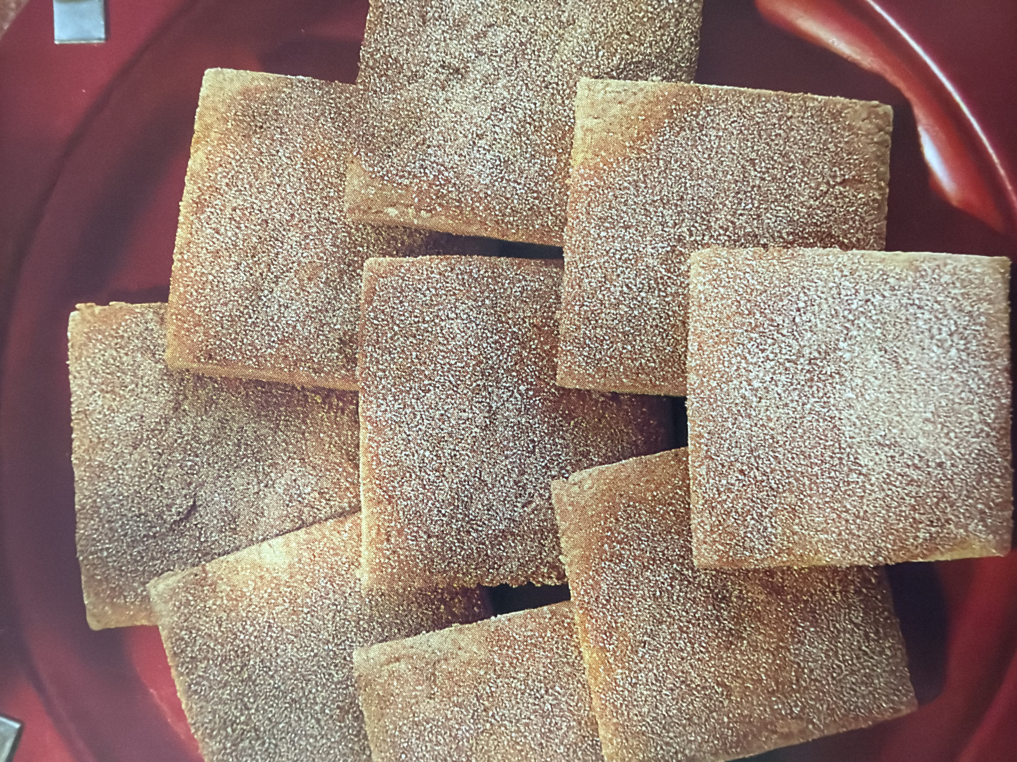 This media file shows a close-up overhead view of several square-shaped cookies or biscuits arranged in a pile. The items are a light brown color and appear to be dusted with a white granular substance, likely powdered sugar or cinnamon sugar. The cookies are stacked on top of each other, with some overlapping. The background is a blurred, deep red color, suggesting the cookies are on a plate or serving dish. There is no visible text. The lighting is consistent and does not indicate a specific time of day or weather conditions. The overall impression is of a food item, likely a baked good, presented for consumption.