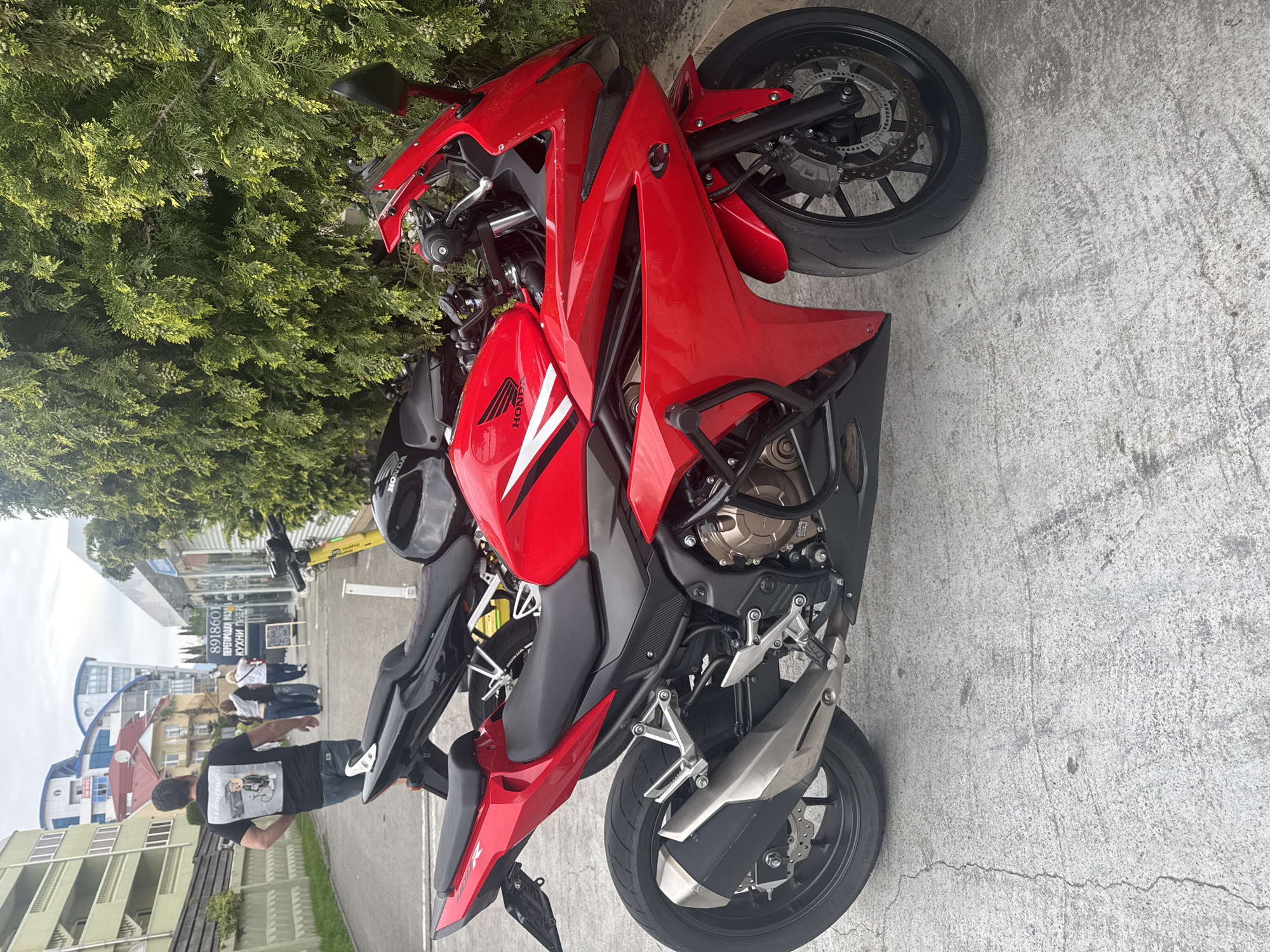 A red Honda motorcycle is parked on a concrete surface next to a bush or tree. In the background, people are walking on a sidewalk in front of buildings. A sign in Russian is visible. Based on the Russian language sign and architectural style, the location is plausibly Sochi, Russia.