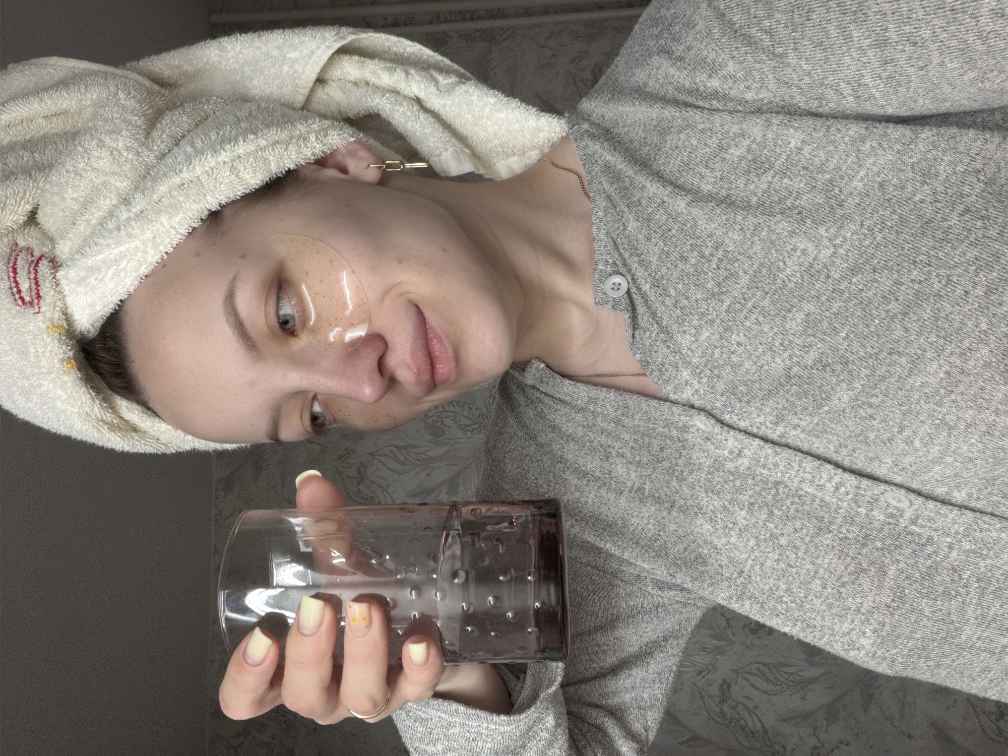 The image shows a woman with a towel wrapped around her head, wearing an eye mask. She is holding a glass with water in it. She is wearing a grey top. The location is not specified, so I cannot confirm the location is Reshetnikovo, Russia.