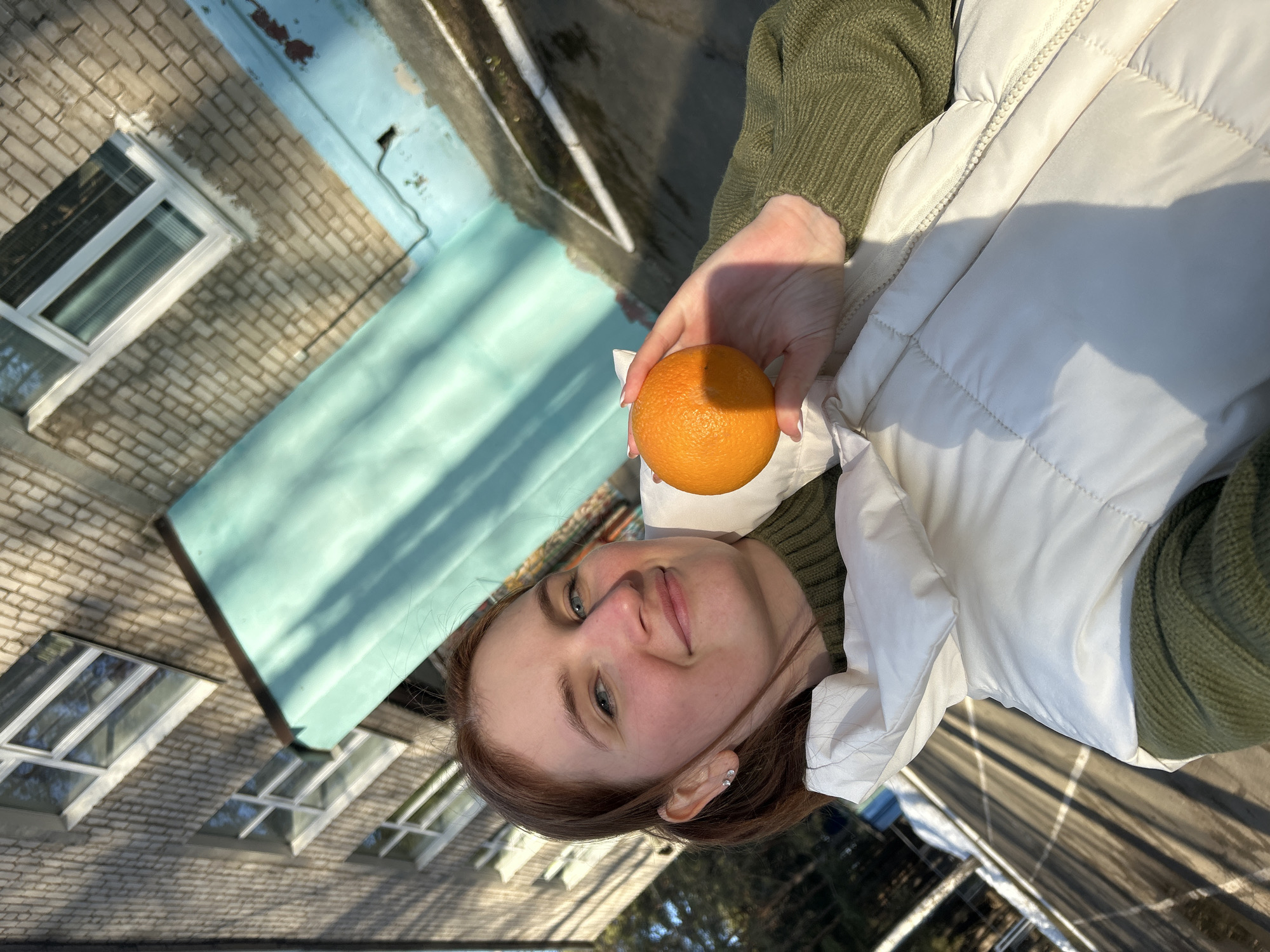 A young woman with auburn hair and light skin holds an orange in her right hand. She wears a green sweater and a white puffy vest. The background reveals a brick building with white-framed windows and a light blue awning. Beyond the building, a road and trees are visible. The location is Chaikovsky, Russia, based on the architectural style typical of the region.