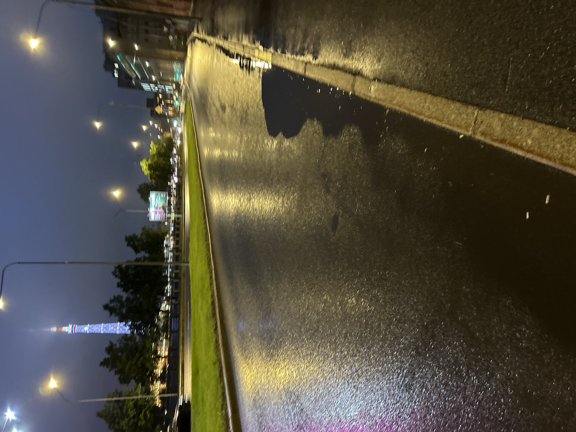 The image depicts a wet asphalt road in a city at dusk or night. The road surface reflects the light from street lamps and distant buildings, creating a glistening effect. The sky is dark blue, with some clouds visible. A tall tower, illuminated with colorful lights, stands prominently in the background, suggesting it is a landmark. Trees line the side of the road, and a patch of green grass is visible to the left. The street appears to be mostly empty, with only a hint of a vehicle on the far left and a distant light that could indicate other traffic. The scene is a street-level perspective looking down the road.