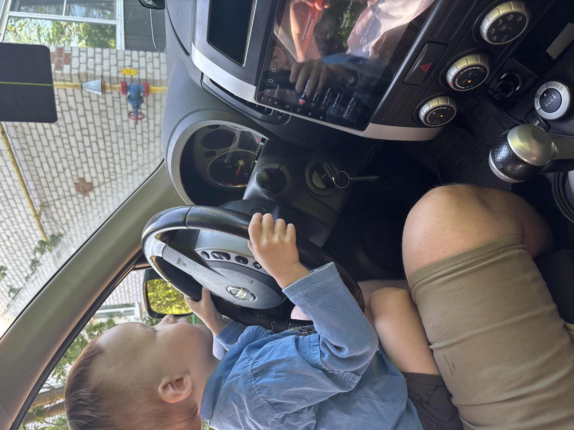 A toddler is sitting in the driver's seat of a car, gripping the steering wheel. The car's dashboard, infotainment screen, and gear shift are visible. An adult's leg is next to the child. The car is parked in front of a building with a yellow gas line and a blue valve on the exterior. The gas infrastructure suggests an urban environment. The infotainment screen has options in Russian. The OCR states a song on the screen by the name of Low feat T-Pain.
The location may be Горный, Russia, based on the metadata.