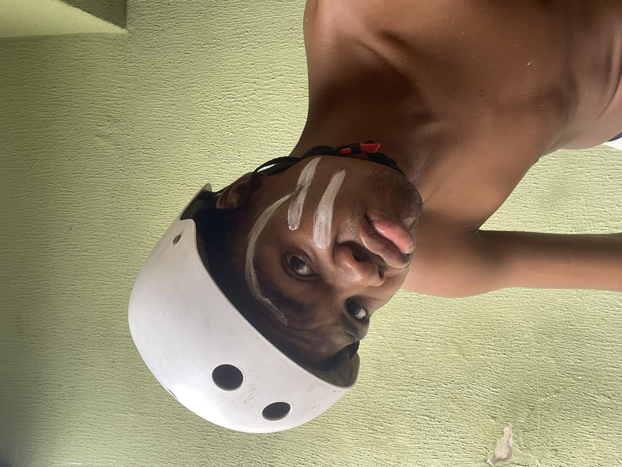 A close-up shot shows a brown-skinned person wearing a white helmet with circular vents. The person has white stripes painted on their forehead and is wearing a black necklace with red accents. The background consists of a light green textured wall. The image appears to have been taken indoors. The location in Ado, Nigeria is not discernible.