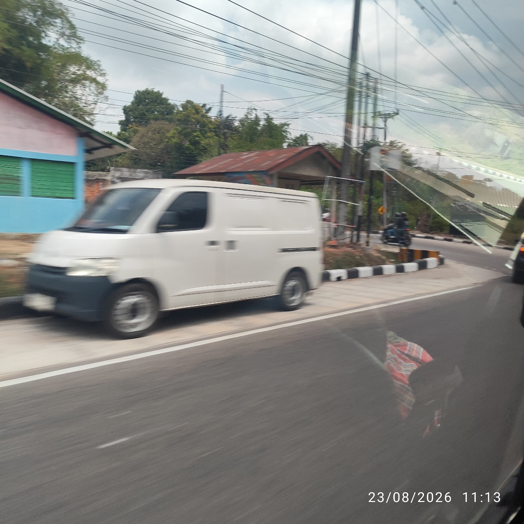 A white cargo van is captured in motion on a paved road under an overcast sky. The van appears to be a Toyota Hiace or similar model, with its front left wheel visible and blurred due to motion. To the left of the road, a light pink building with a blue trim and green shutters is partially visible, along with trees and some overhead power lines. Further down the road, a building with a rusty tin roof is present. In the distance, a black motorcycle with two riders is seen navigating the road. The scene is marked with a black and white striped barrier along the side of the road. The image is dated August 23, 2026, at 11:13 AM.