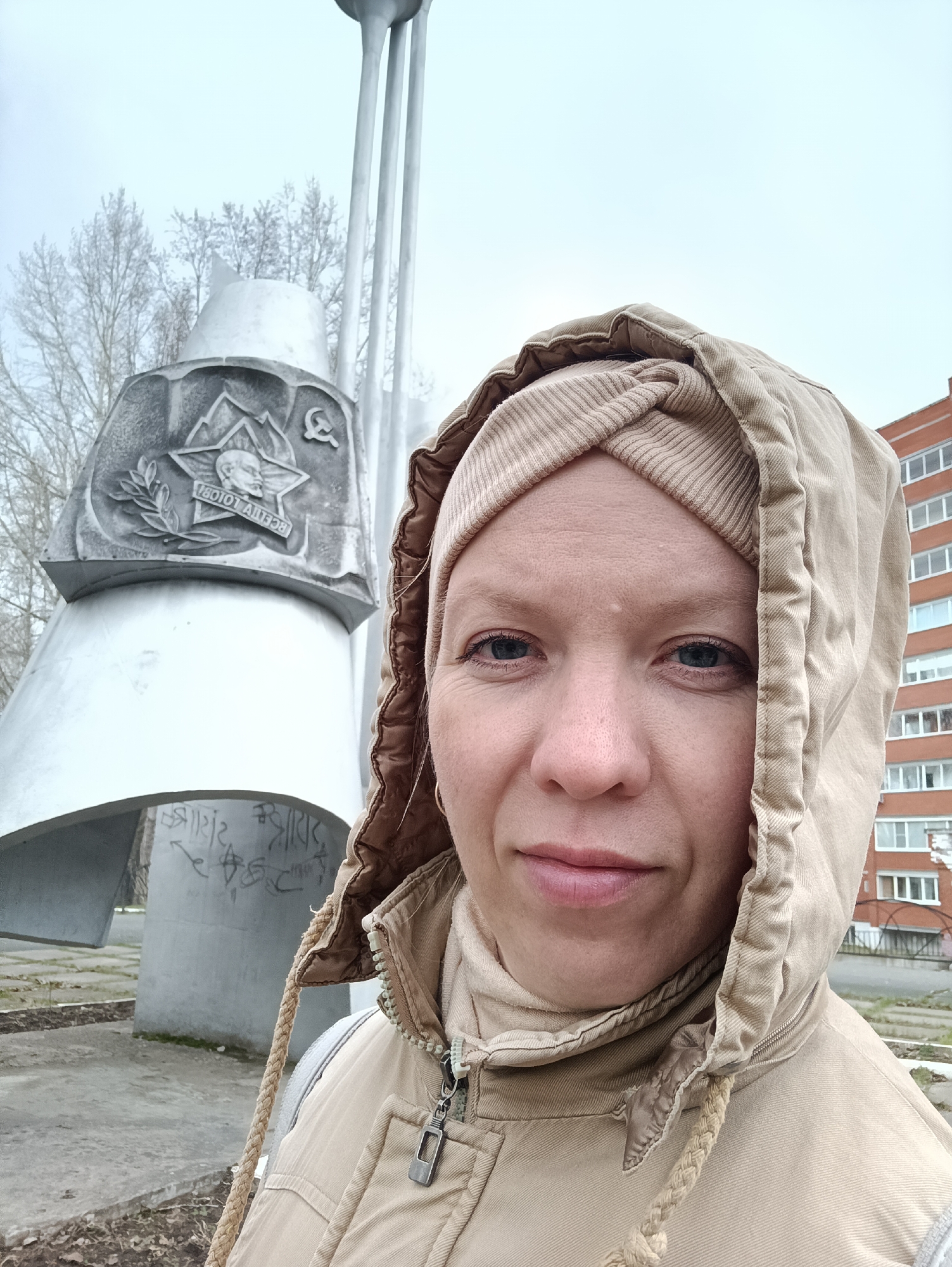 A woman is captured in a close-up selfie, standing outdoors in Chaikovsky, Russia, on an overcast day. She is in the immediate foreground, looking directly at the camera with blue eyes and a neutral to slightly smiling expression. She wears a beige hooded jacket with the hood up and a textured, beige head covering beneath it.
Behind her to the left, a large, light-colored concrete or stone monument dominates the middle ground. The monument features a prominent relief carving displaying a five-pointed star. Within the star, a profile bust of a man (likely Vladimir Lenin) is depicted, accompanied by a laurel branch on the left and a hammer and sickle emblem on the right. Below the bust, the Russian text "ВСЕГДА ГОТОВ!" (ALWAYS READY!) is inscribed. The base of the monument is adorned with some graffiti.
Further in the background, a tall, slender, multi-pronged structure rises towards the sky. On the left side, bare trees indicate a cool season, while on the right, a multi-story red-brick building with several windows is visible. The ground beneath the monument is paved with concrete and shows some scattered debris. The overall setting suggests a public space or park, likely featuring a Soviet-era memorial.