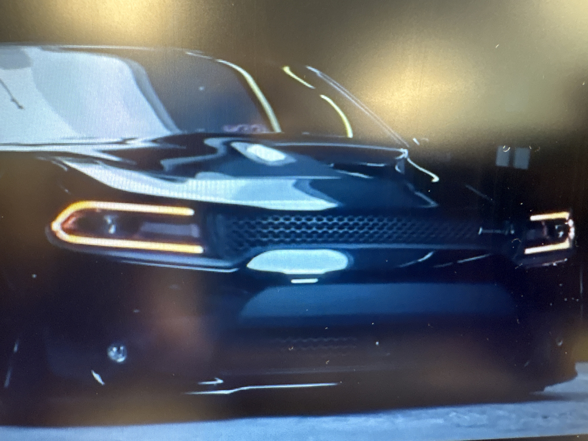 The image shows a close-up, slightly blurry, frontal view of a black car, likely a Dodge Charger, parked on an asphalt surface. The car has its headlights illuminated, revealing a distinct horizontal LED strip within the headlight assembly. The grille of the car is visible, featuring a mesh pattern. A portion of the windshield and hood is also captured. The background is dark and out of focus, with some indistinct shapes that could be buildings or other objects. There are no people or any discernible city features that would indicate Sofia, Bulgaria, or any specific urban environment. The overall impression is that of a vehicle photographed in low light conditions, possibly at night.